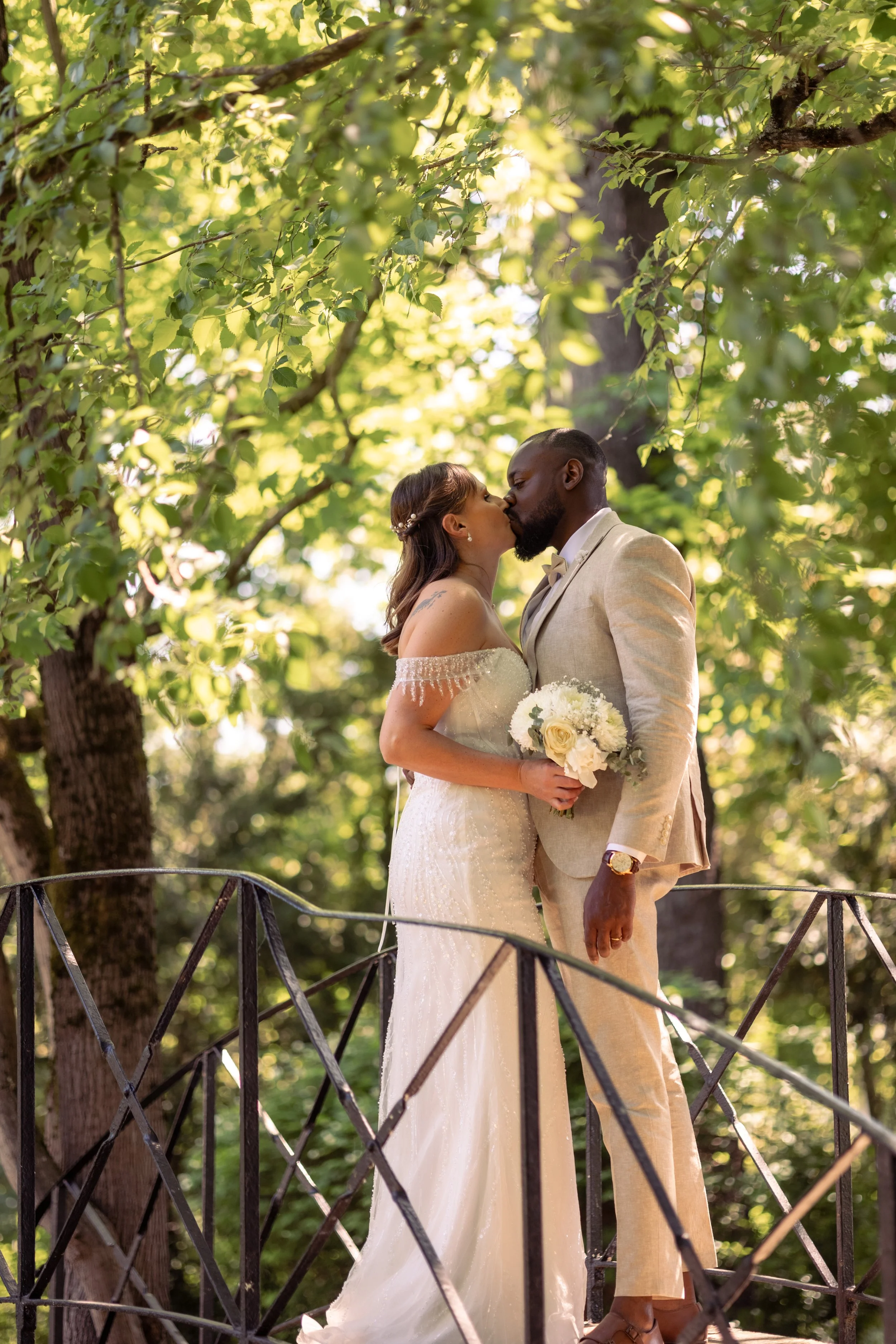 Un couple en mariage s'embrassant sur une petite terrasse en plein air, entourés d'arbres verts, avec la femme tenant un bouquet blanc, vêtus d'élégantes tenues de mariage dans une lumière douce et naturelle.