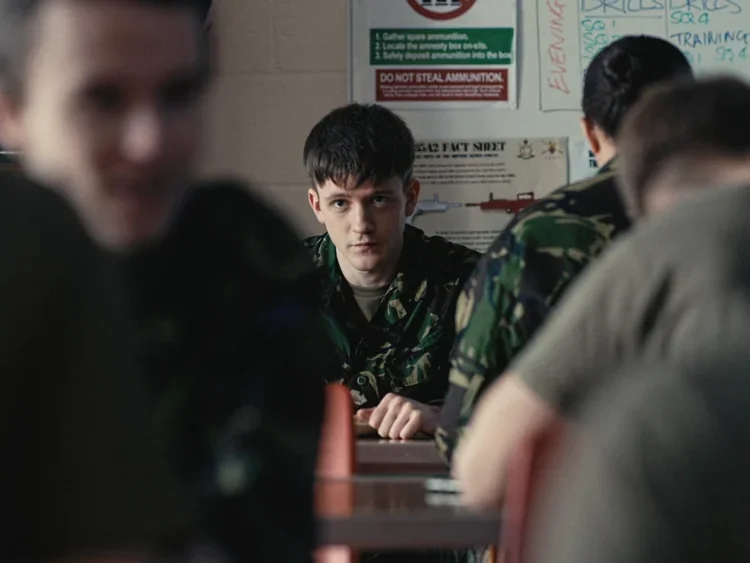 A young man in military camouflage uniform looking directly at the camera while sitting at a table in a classroom or briefing room with other people, some with similar camouflage uniforms and some in civilian clothing, with a whiteboard and informational posters on the wall behind him.
