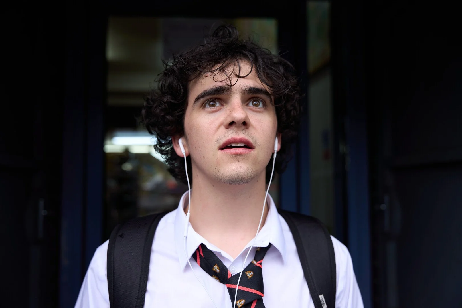 A young man with curly dark hair, wearing a white shirt, black backpack, and a black tie with red and white pattern, standing outdoors with a surprised or concerned expression, listening to white earbuds.