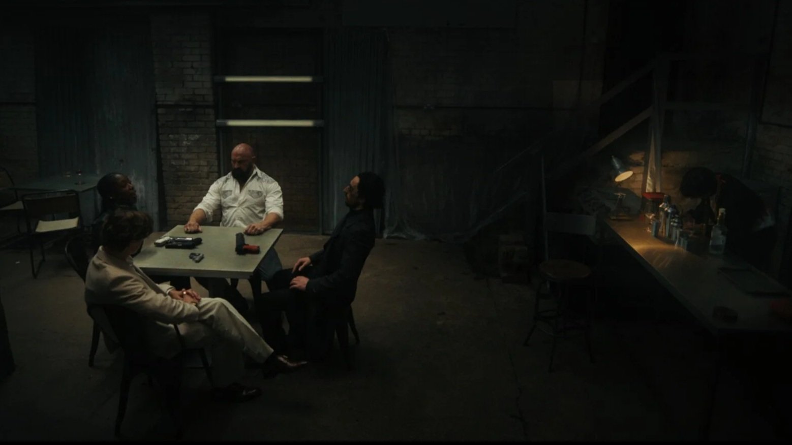 Four people sitting at a table in a dark, industrial-style room, with guns on the table, and a bartender working behind a bar with drinks.