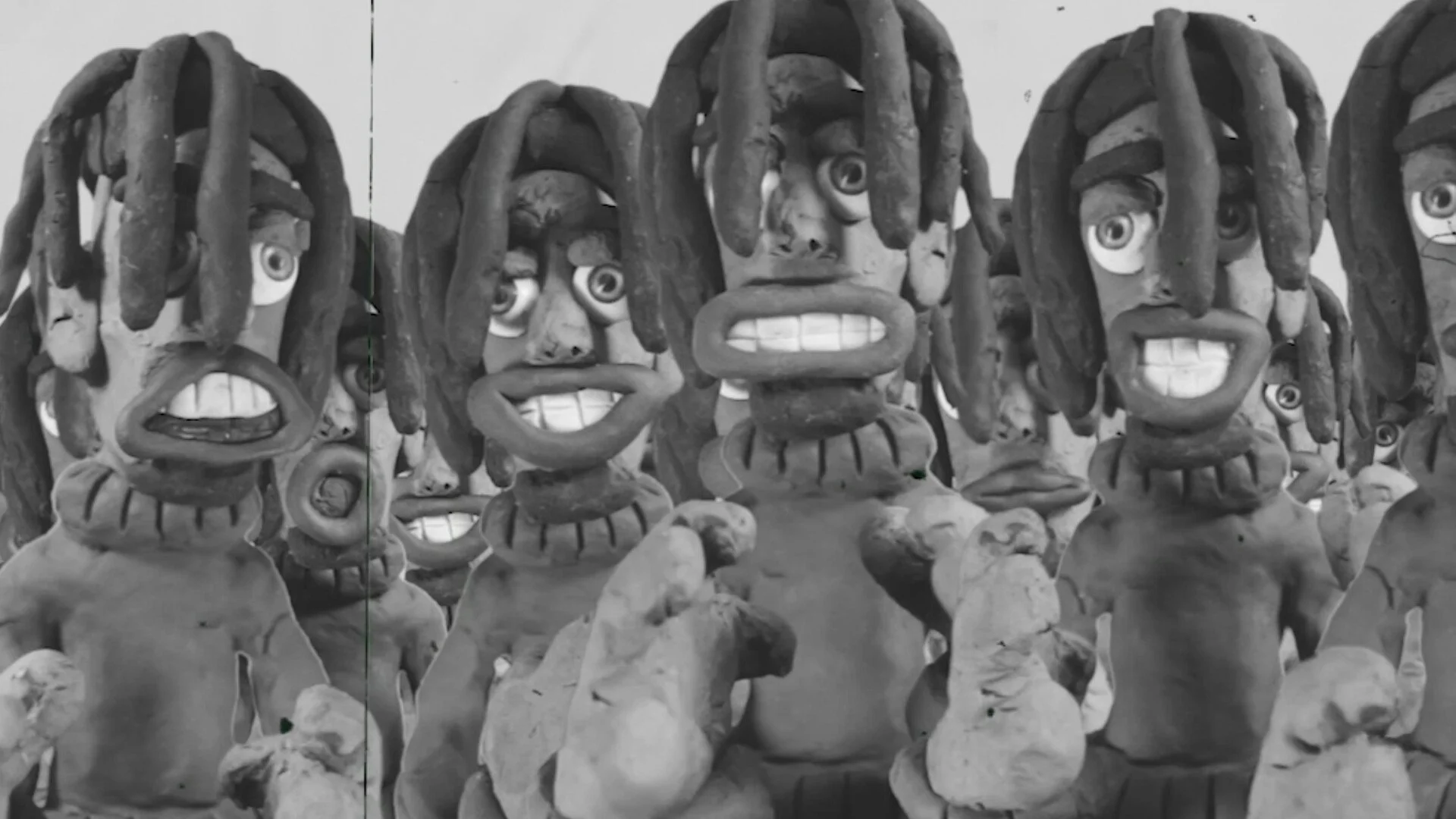A group of cartoonish clay sculptures of human faces with large eyes and dreadlocks, some showing teeth, arranged in a statuesque formation.