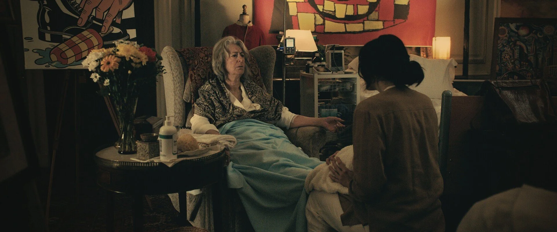 An elderly woman lying in bed holding hands with a caregiver in a cozy, dimly lit room decorated with colorful artwork and flowers.