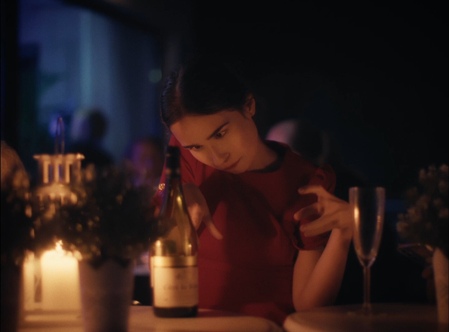 A young woman in a red dress sitting at a table with bottles and glasses, looking down thoughtfully in a dimly lit setting.