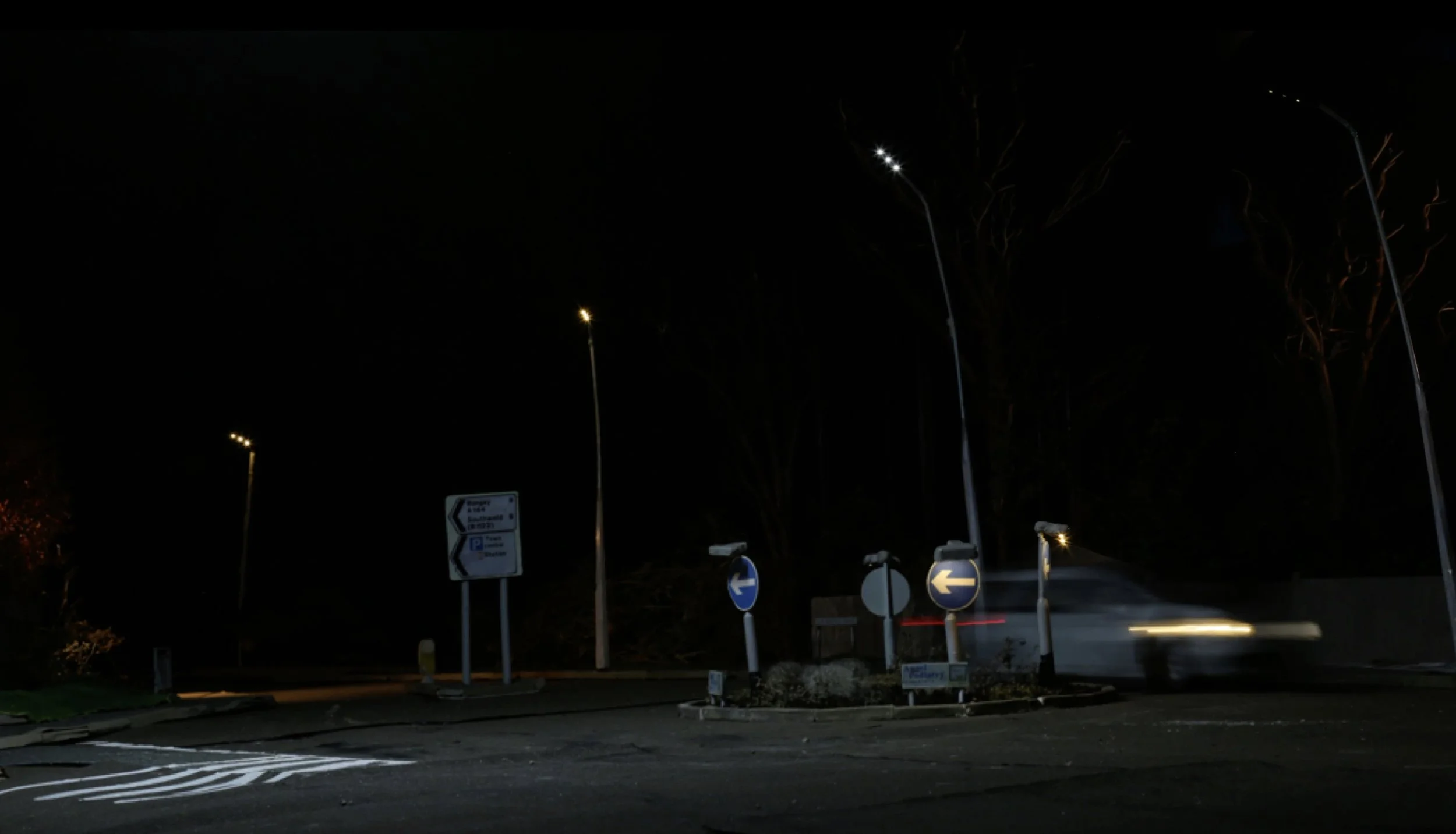 Nighttime scene of a roundabout with directional signs and streetlights, with a blurred white car moving through.