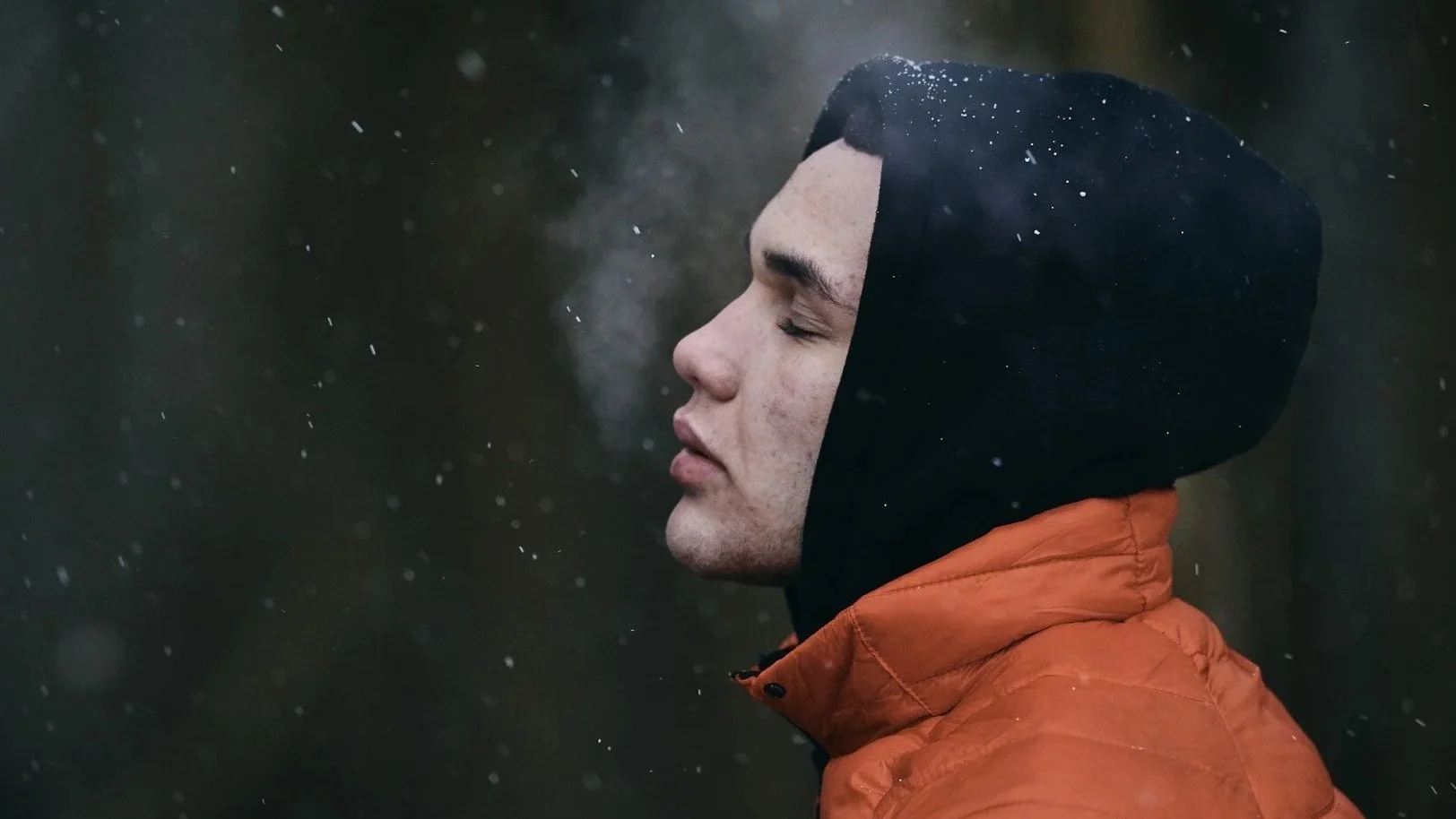 A young man in a black hoodie and orange jacket stands in the rain with his eyes closed, facing the side, with water droplets falling around him.