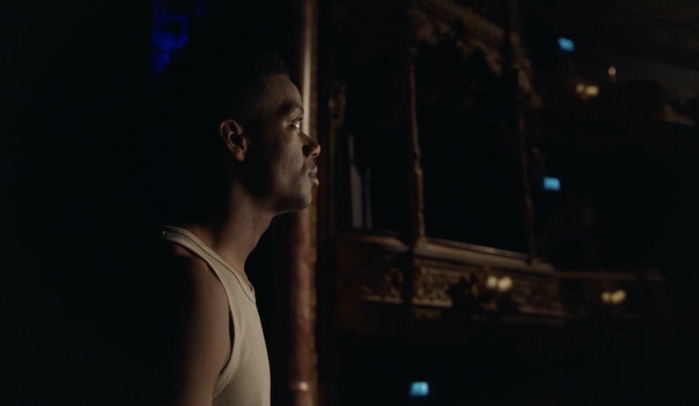 Side view of a young man with dark hair and a beige tank top, standing in a dark room with a decorative balcony or railing in the background.