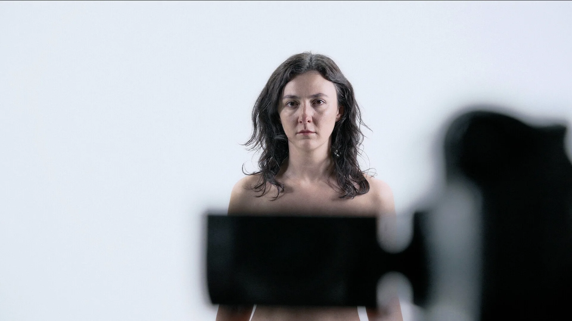 A woman with shoulder-length wavy dark hair standing against a plain white background, looking directly at the camera with a serious expression.