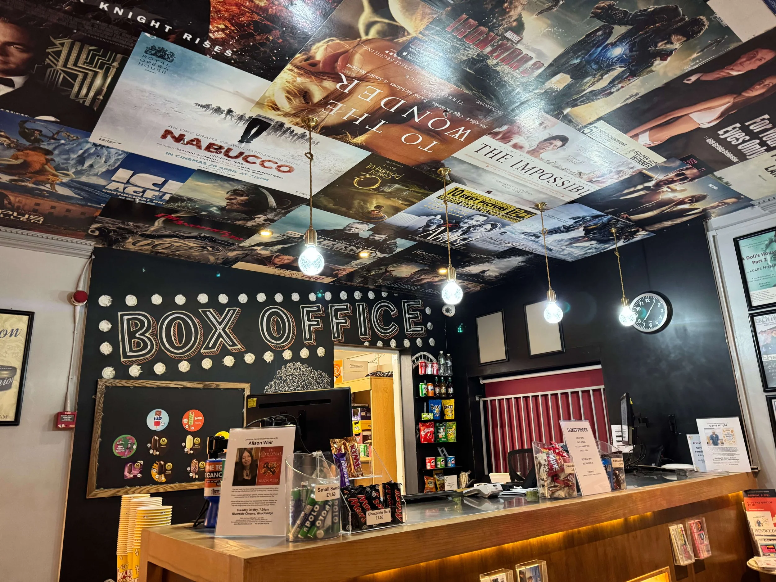 View of a box office area with a black wall and a chalkboard sign reading 'BOX OFFICE' in white letters with decorative dots. The ceiling is covered with movie posters. There are hanging light bulbs, a clock, and a counter with tickets, candies, and promotional materials.