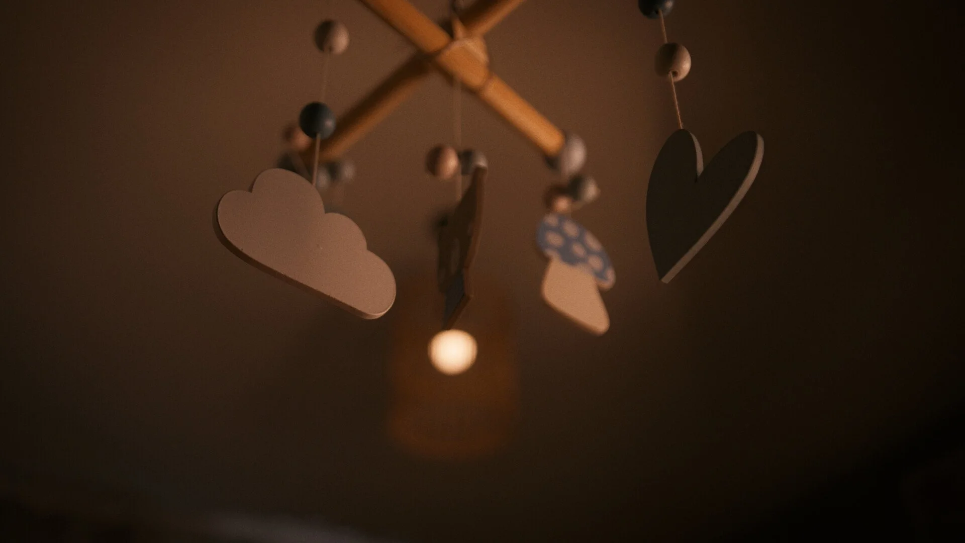 Dark photo of a hanging mobile with cloud, heart, and star shapes, illuminated by a warm light.