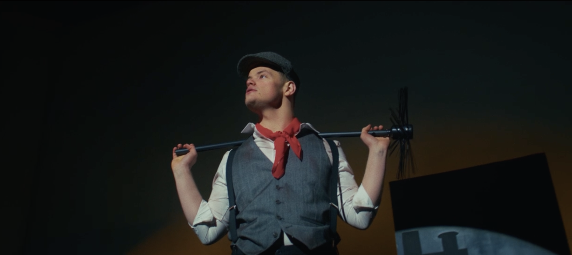 A man dressed as a rebellious farm boy from the 1940s or 1950s, wearing a flat cap, vest, and red neckerchief, holding a rake over his shoulders, looking confidently into the distance, with a dark background and a windmill in the distance.