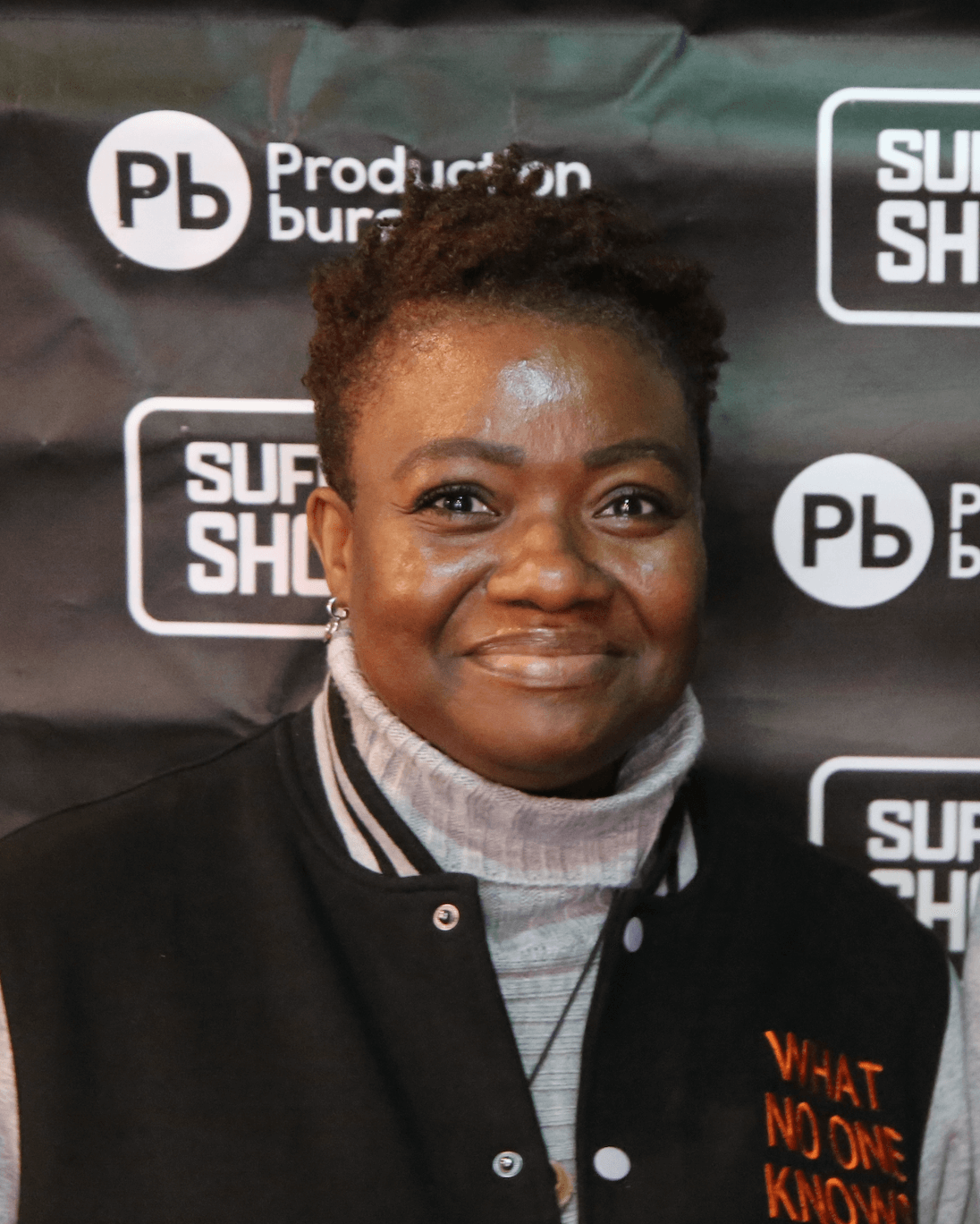 A woman with curly black hair wearing a patterned blouse and a gold necklace, smiling in front of an abstract painting.
