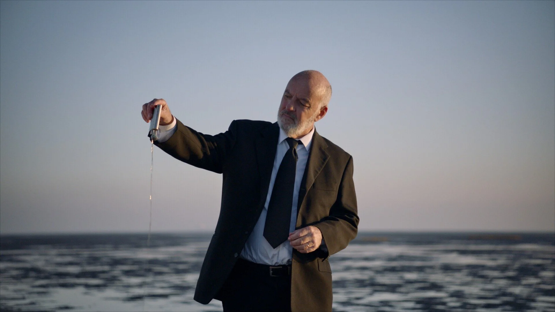 A man in a black suit, white shirt, and black tie stands on the beach at sunset, holding a test tube with a liquid.