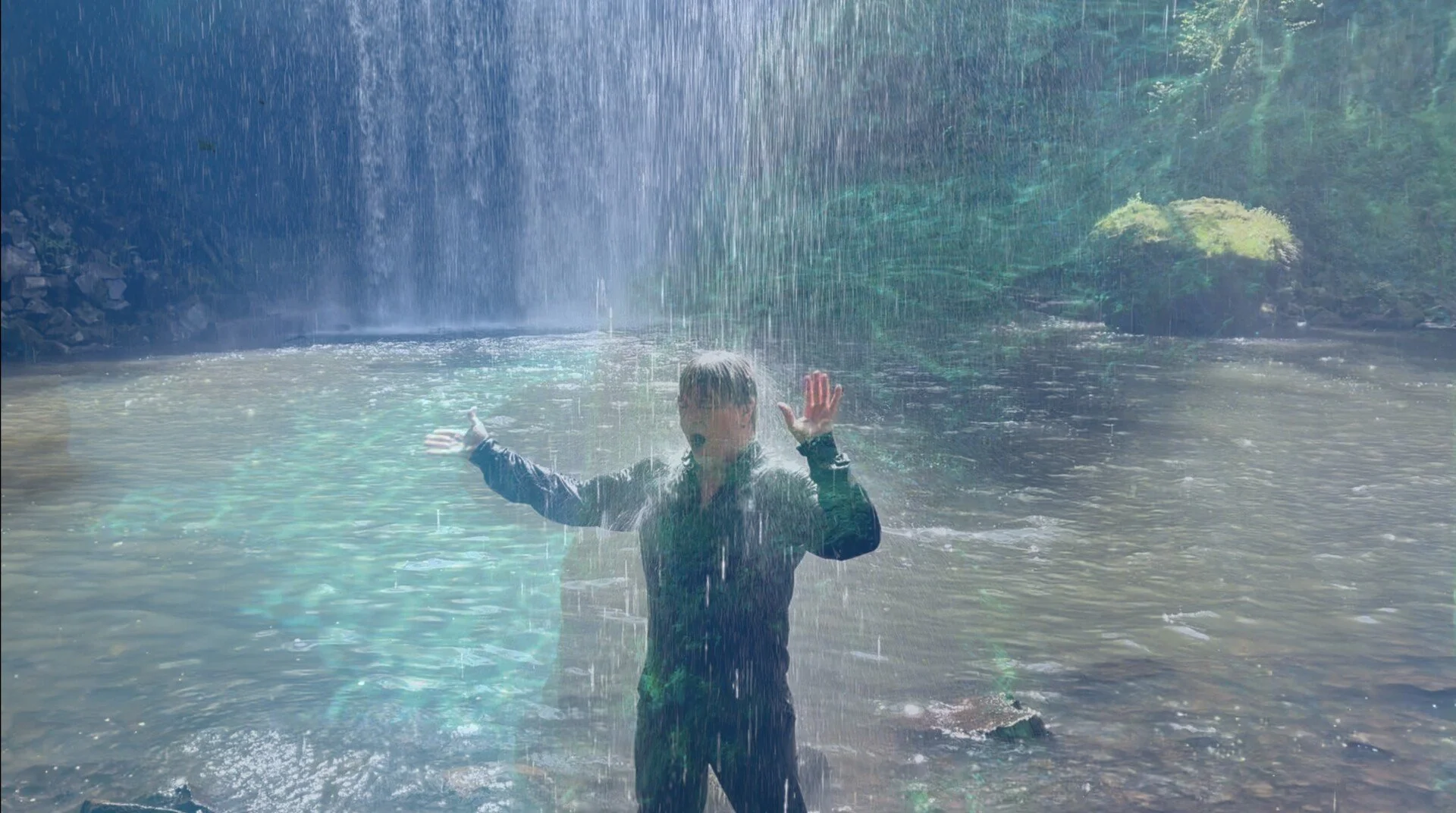 A person standing under a waterfall in a natural setting, arms outstretched, with water cascading from above.