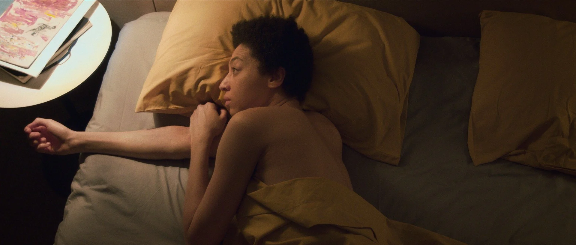 A woman lies on her side in bed with yellow pillows, appearing to be sleeping or resting, next to a bedside table with magazines and a fork on top.