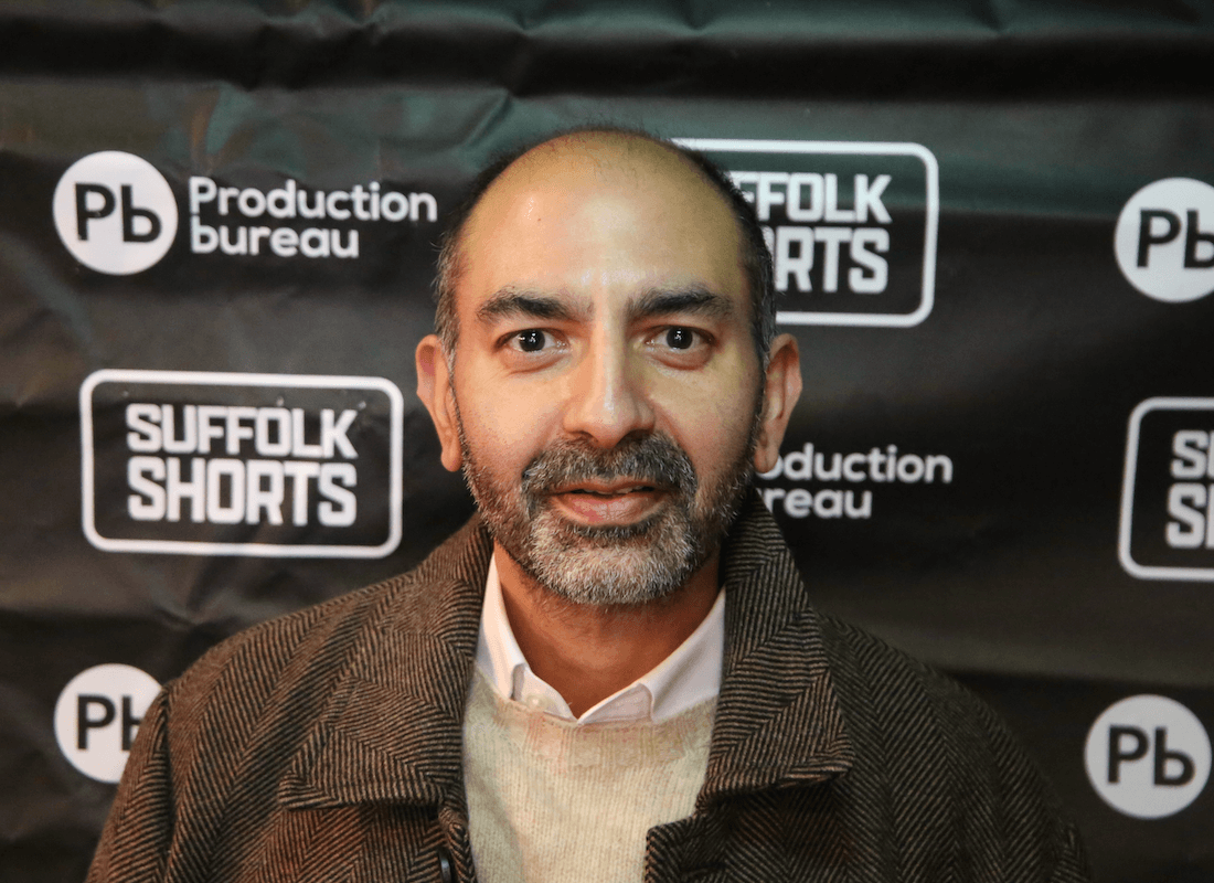 Close-up of a man wearing a gray hat and a navy sweater, inside a room with a wooden bookshelf filled with books in the background.