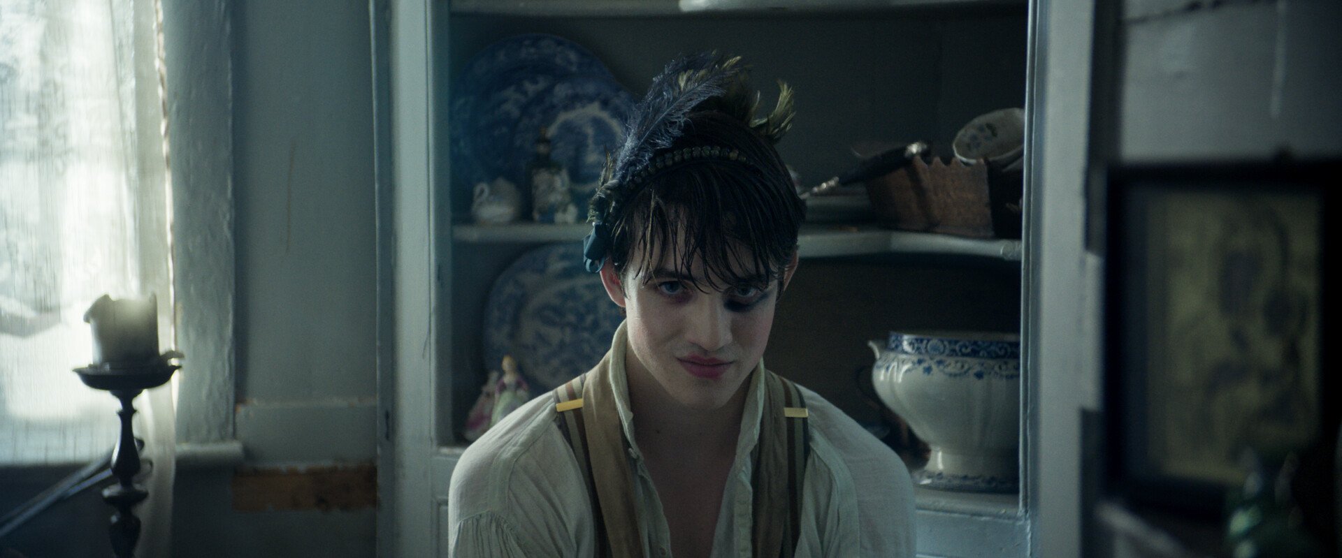 A person with dark wet hair, wearing a white shirt with suspenders, smiling slightly, standing in a dimly lit kitchen with a cabinet in the background.