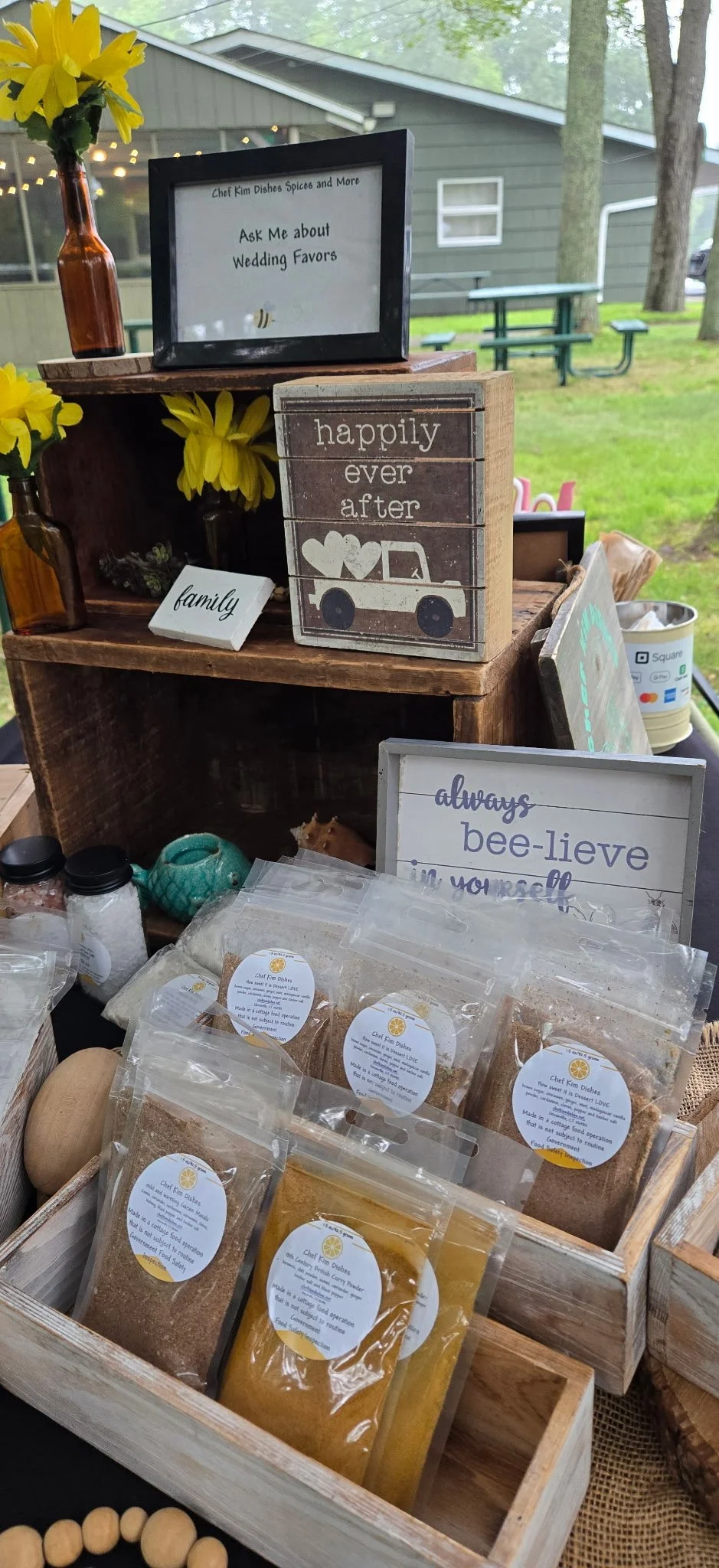 Display of various food seasonings in clear plastic bags, decorative signs with messages like 'happily ever after' and 'always bee-lieve in yourself,' yellow flowers in brown glass bottles, and a small framed sign asking about wedding favors, set outdoors with trees and a green building in the background.