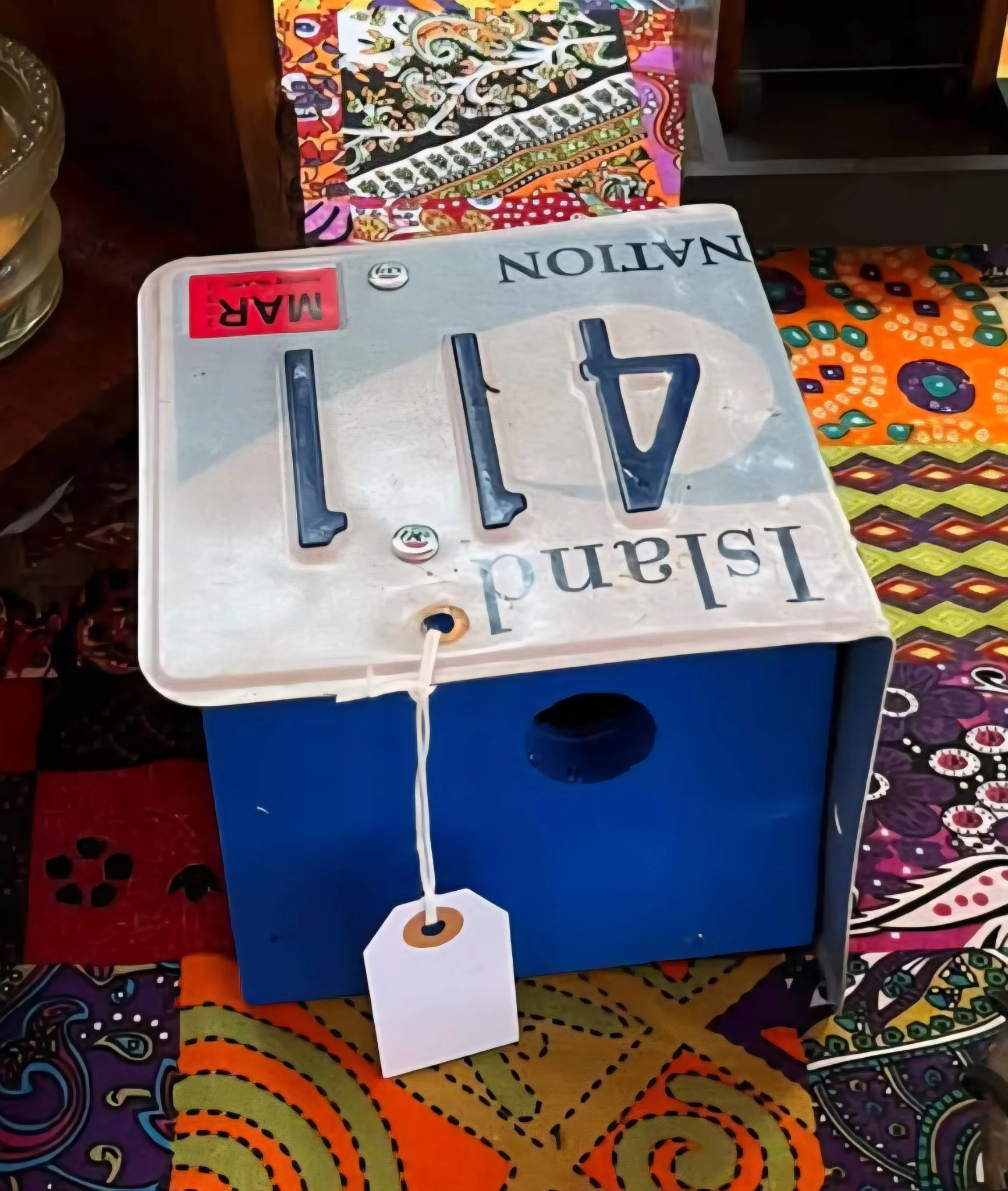 A decorative box with a vintage-style sign on top that reads 'Island Navigation.' The sign has large black letters and a red 'MAR' sticker. The box is placed on a colorful, patterned fabric surface.