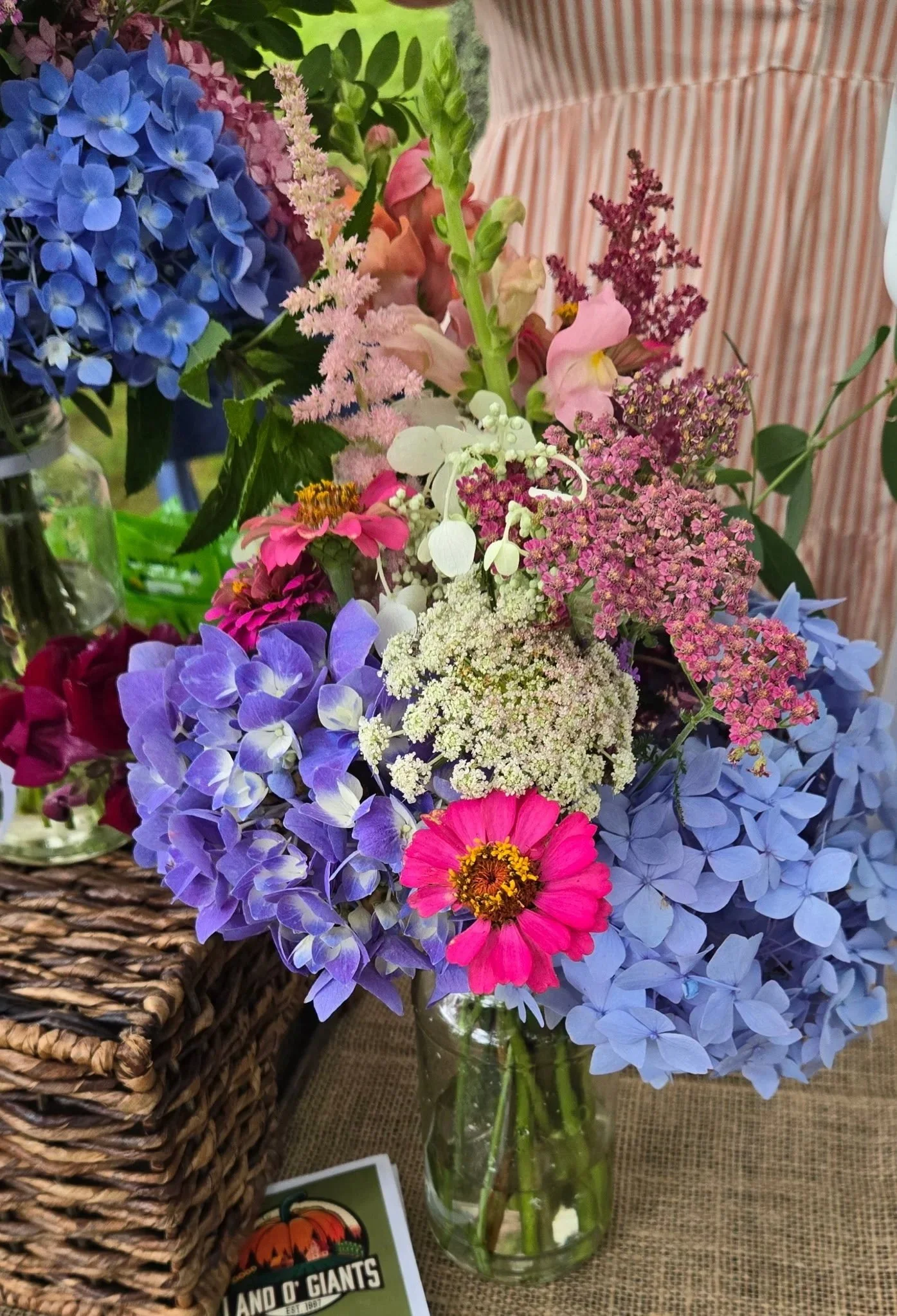 A colorful bouquet of flowers in a glass jar includes blue hydrangeas, pink zinnias, white Queen Anne's lace, pink snapdragons, purple veronica, and other mixed flowers arranged on a table with a woven basket nearby.
