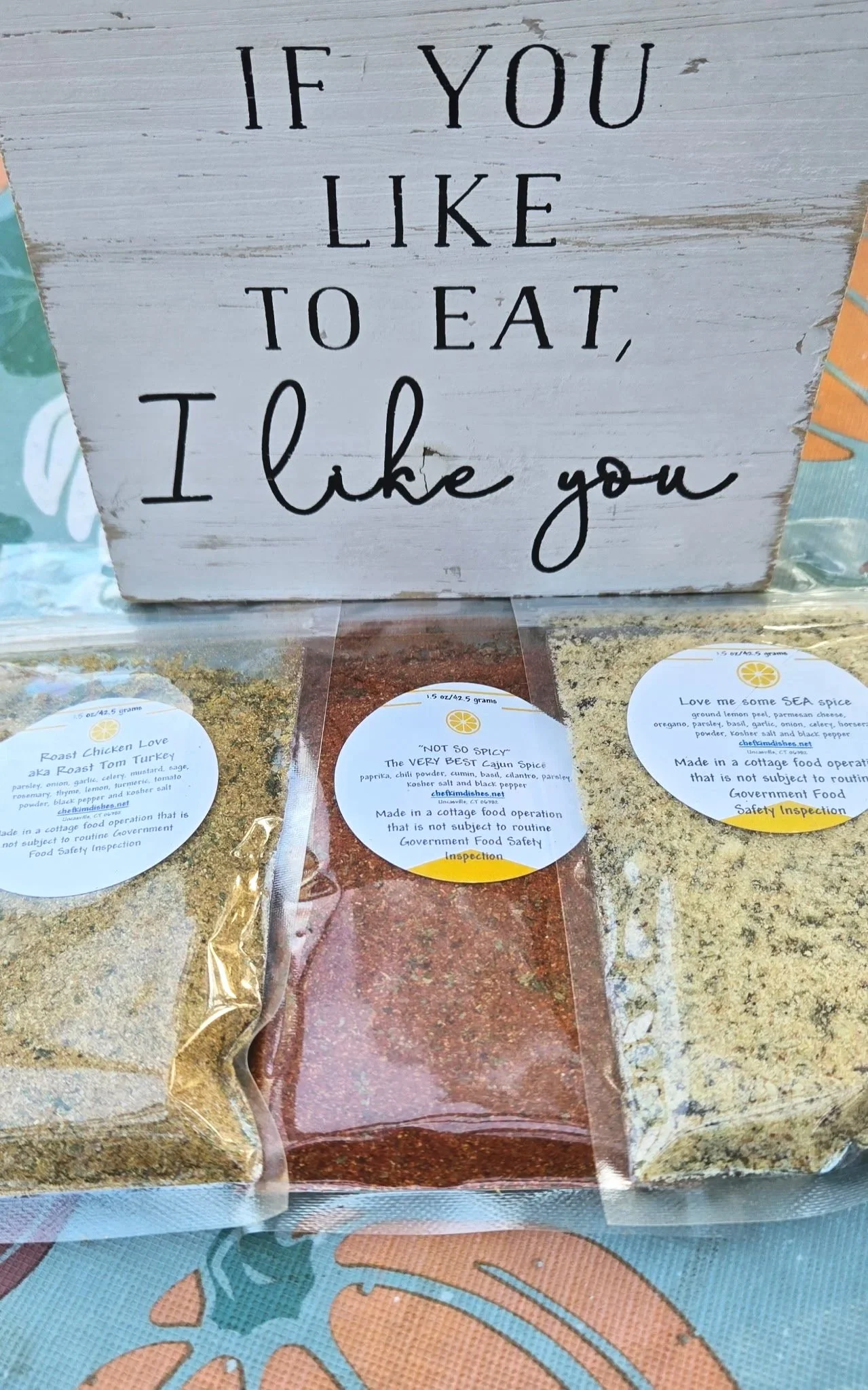 A wooden sign with the quote "If you like to eat, I like you" displayed above three packets of spices with labels indicating their contents and safety inspection stamps.