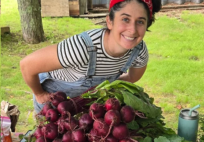 A woman smiling in a striped shirt and overalls leaning over a large pile of freshly harvested red beets with green leaves, outdoors on a grassy area.