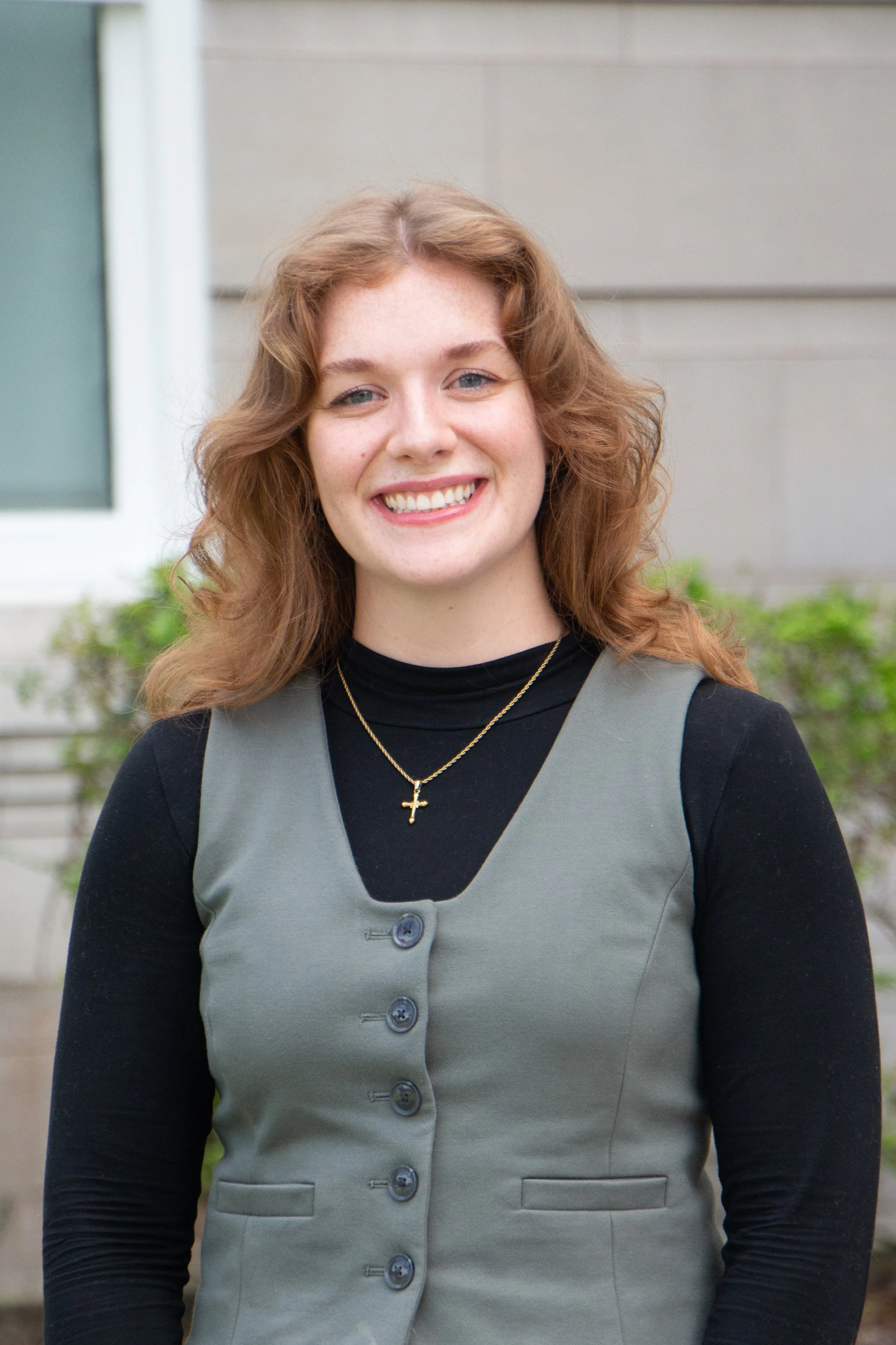 A young woman with red wavy hair, smiling, wearing a black long-sleeve shirt, a grey vest with buttons, and a gold cross necklace, standing outside in front of a building with greenery.