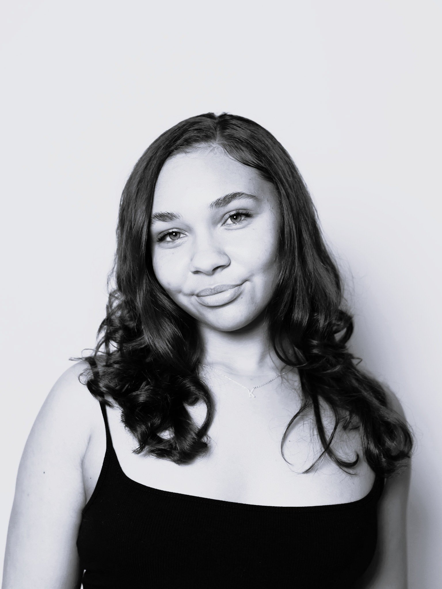 Black and white portrait of a young woman with long curly hair, wearing a black sleeveless top and a delicate necklace, smiling softly against a plain background.