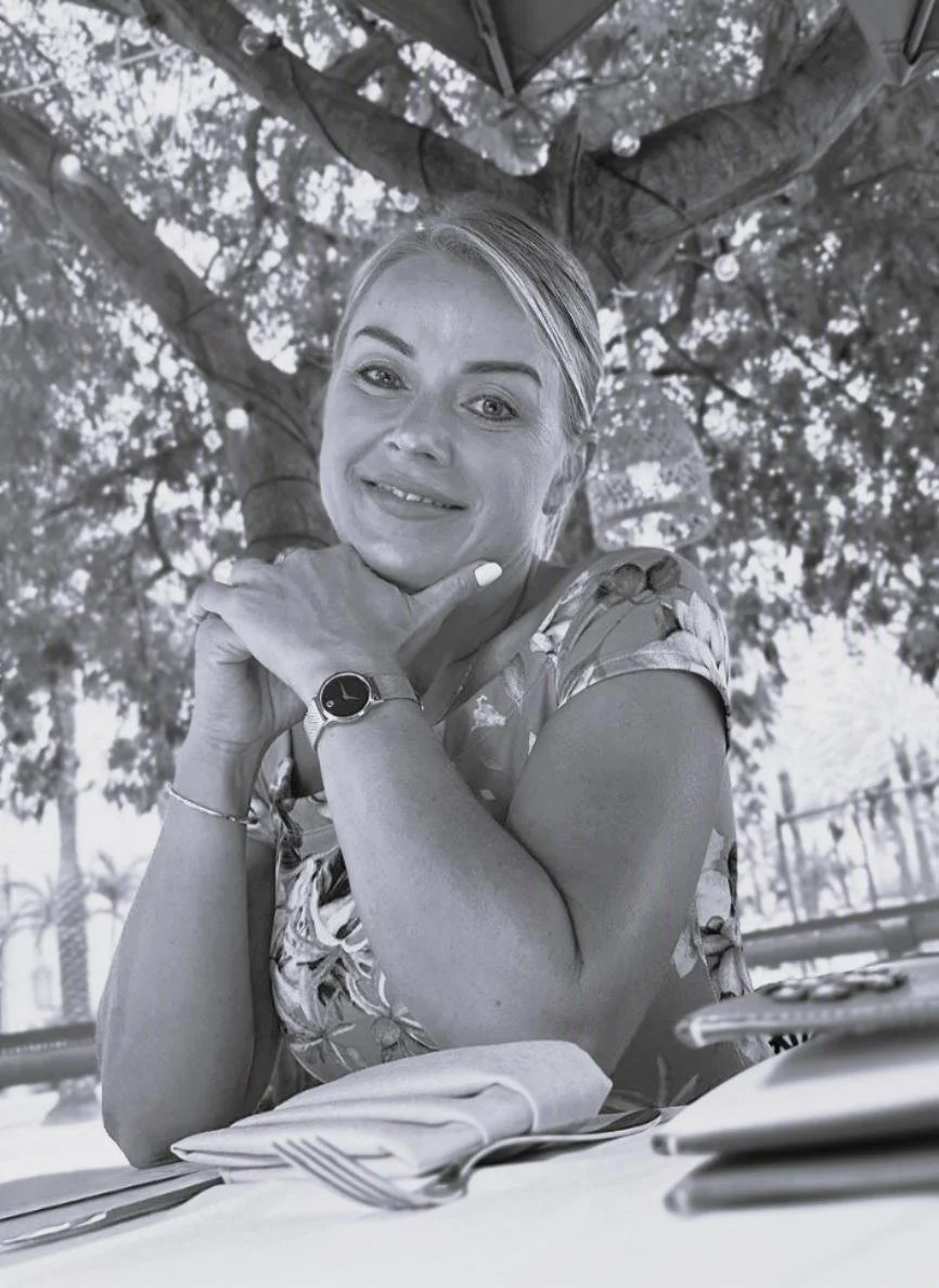 A woman sitting at a table outdoors under a tree, smiling and resting her chin on her hand, wearing a patterned top and a watch on her left wrist.