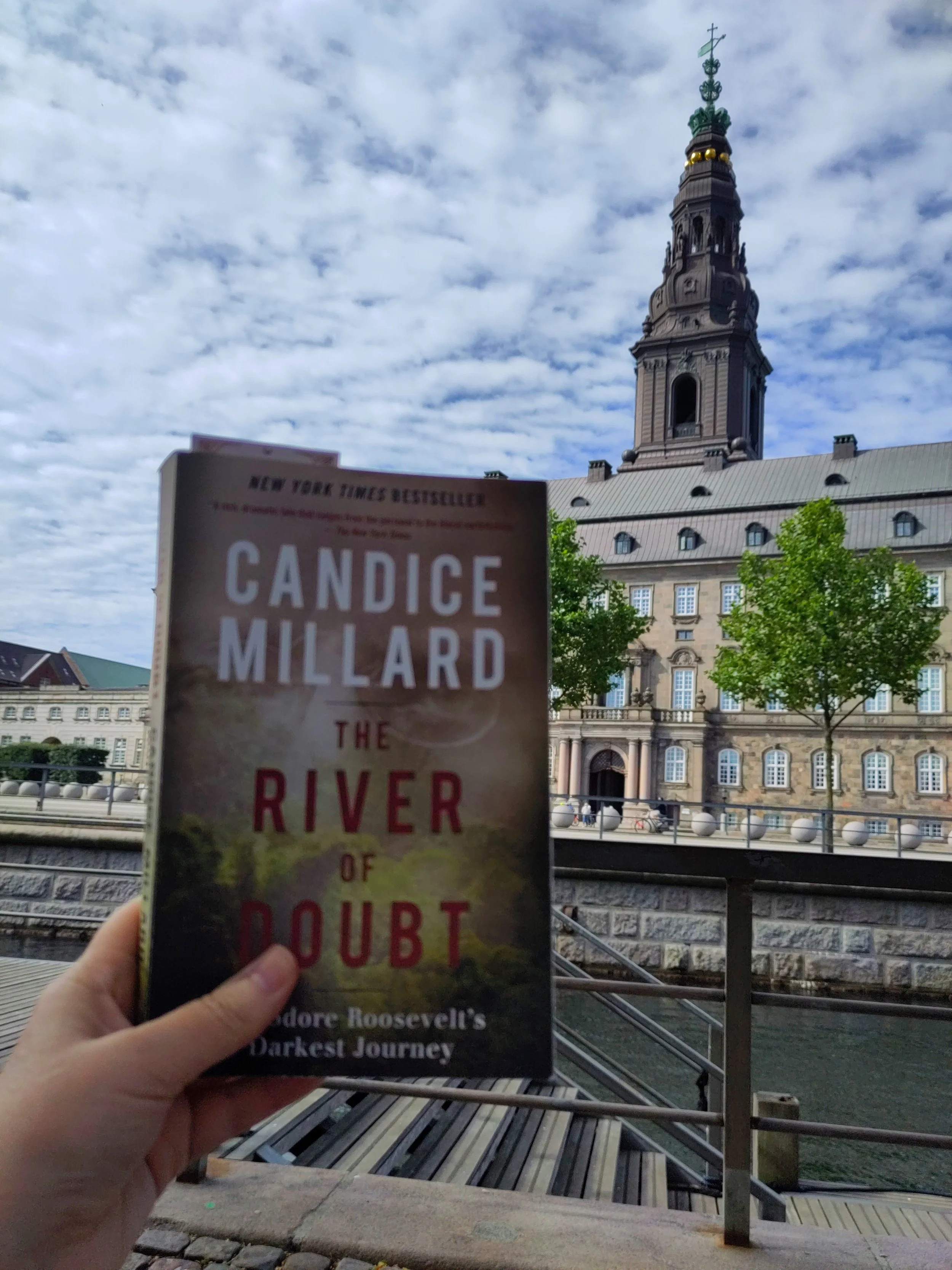A person holding a copy of the book 'The River of Doubt' by Candice Millard with a historic building and a church steeple in the background.