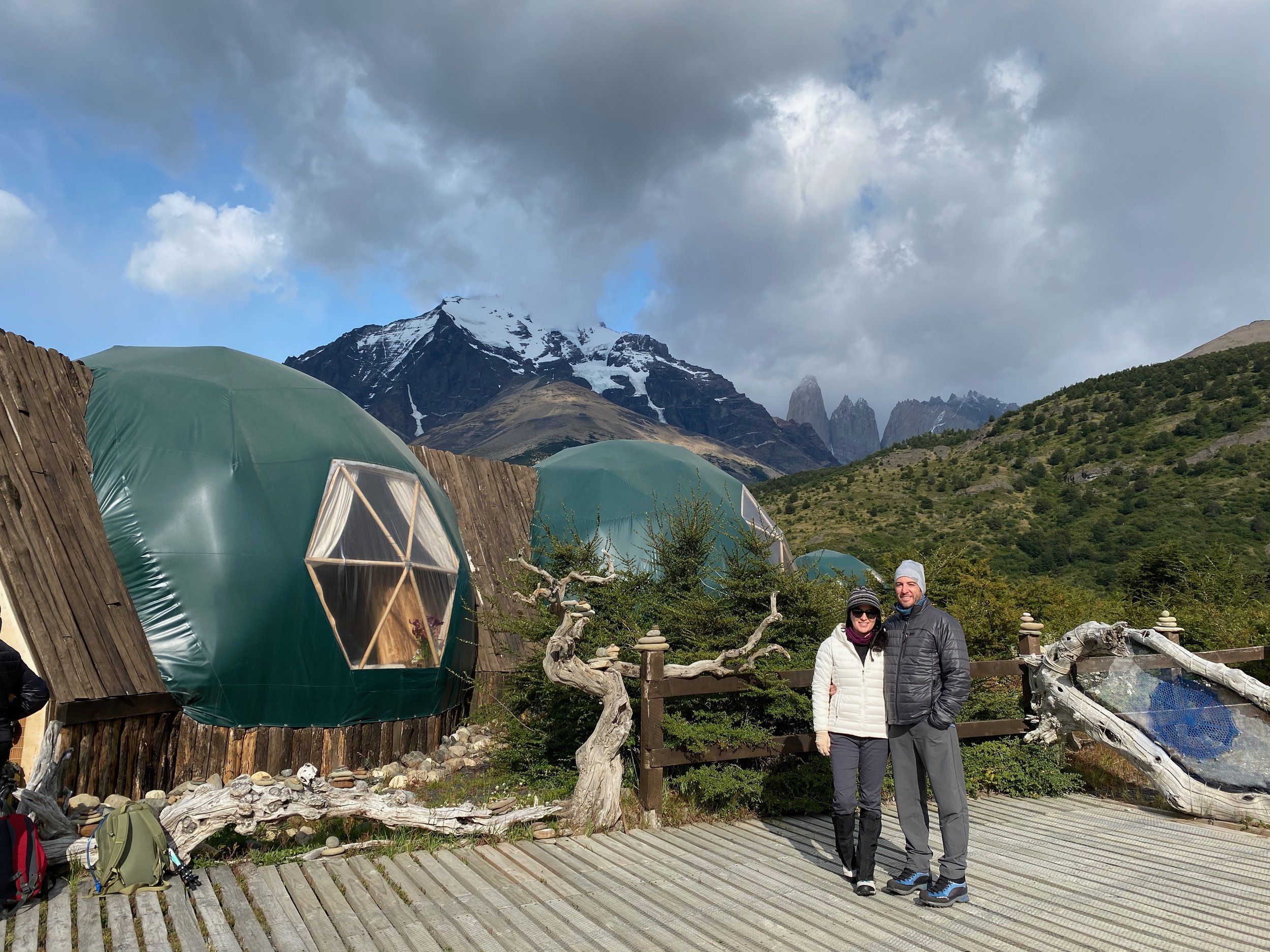Two people standing on a wooden deck in front of eco-friendly dome-shaped cabins with a mountainous landscape in the background.