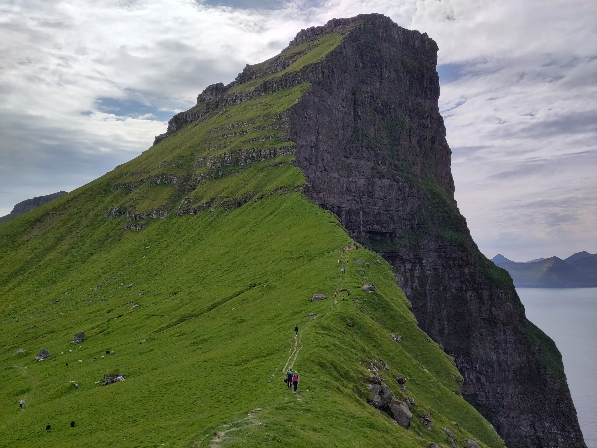 Lush green mountain with steep, layered slopes and a cliff face, with hikers walking along a narrow trail on the mountain's side, overlooking water and distant mountains.
