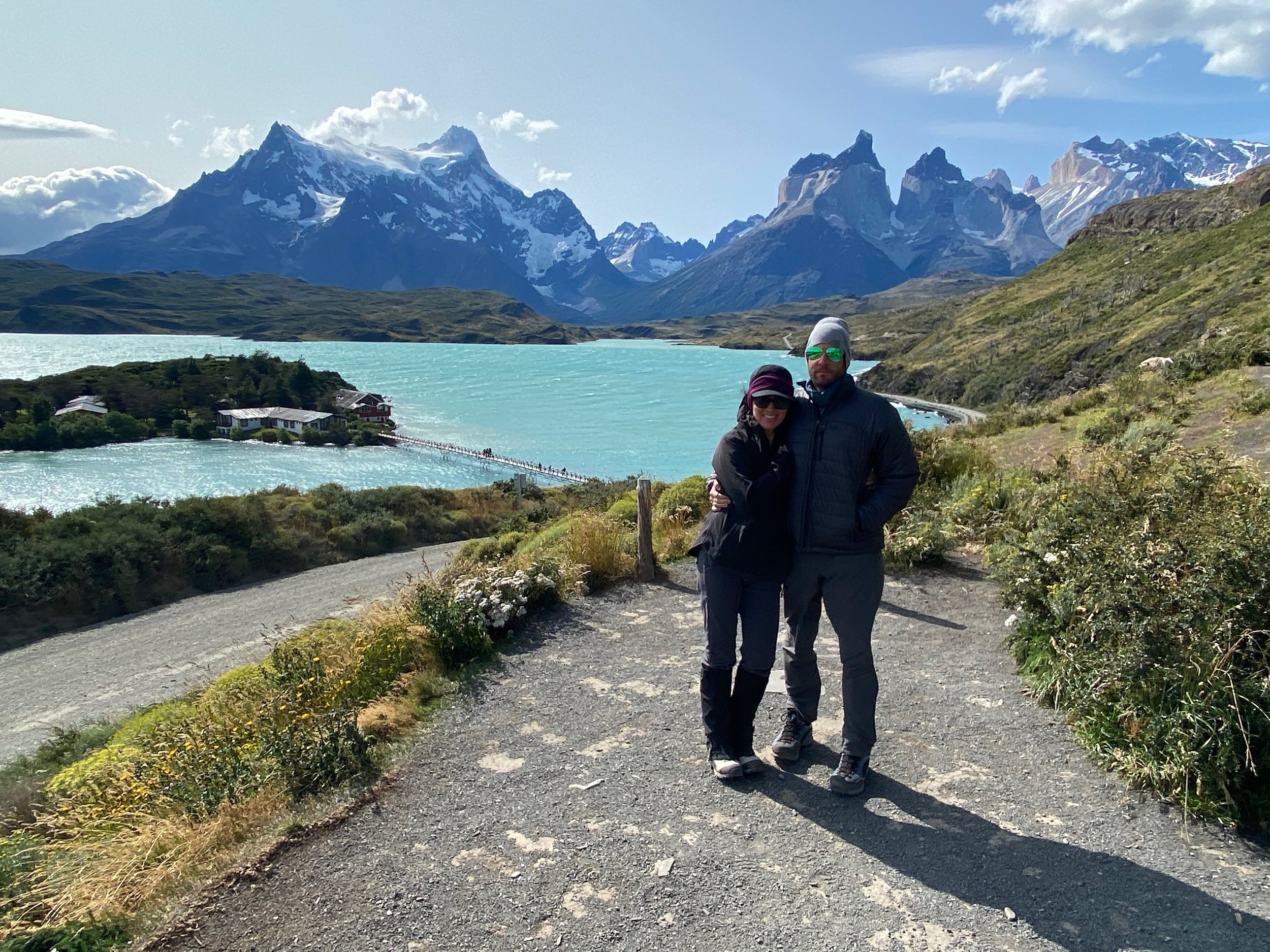 A couple stands on a scenic path with views of turquoise waters and snow-capped mountains under a clear sky.