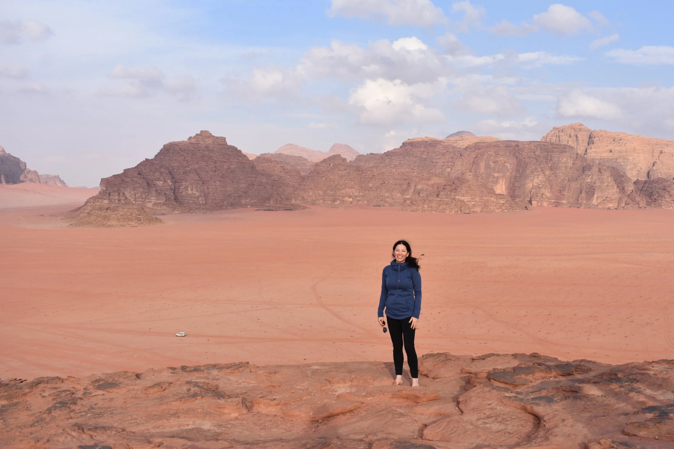 A woman standing on a rocky ledge in a desert landscape with reddish sand, towering sandstone mountains in the background, and a partly cloudy sky.