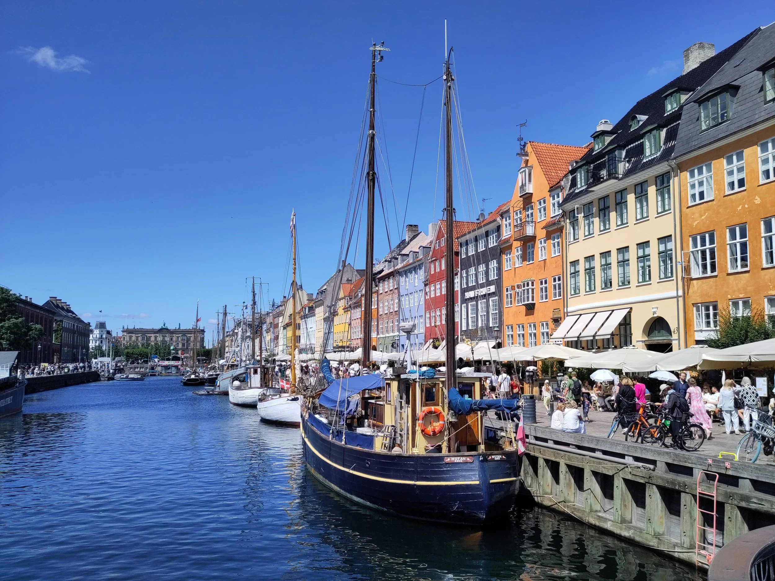A scenic harbor with colorful buildings lining the waterfront, boats docked along the water, and people enjoying the outdoor cafes on a sunny day.
