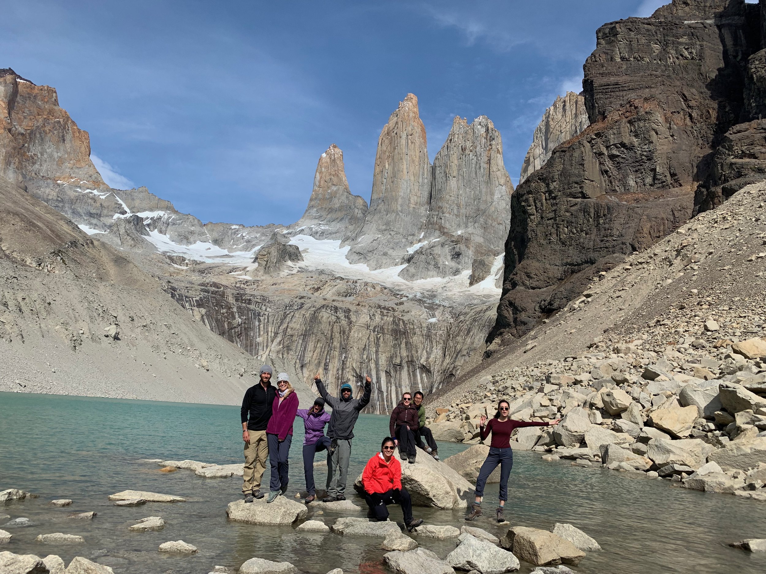 A group of people posing on rocks near a turquoise lake in a mountainous area, with three prominent, jagged granite peaks in the background under a clear blue sky.