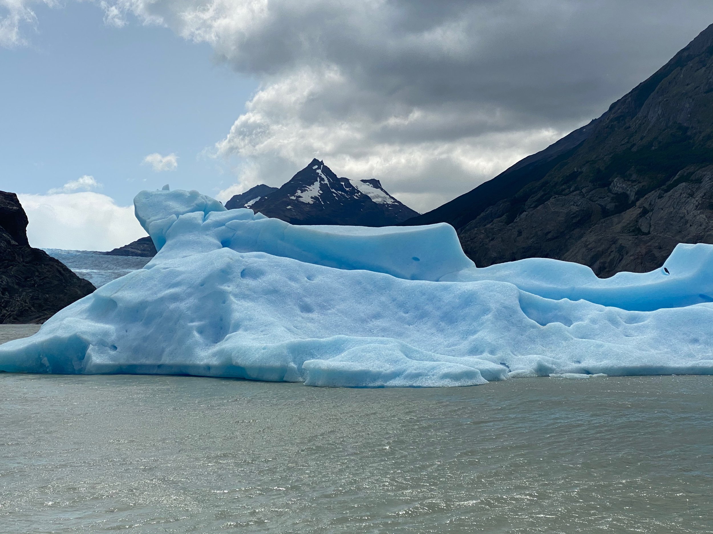 Large blue iceberg floating in a body of water, with mountainous terrain in the background under a cloudy sky.