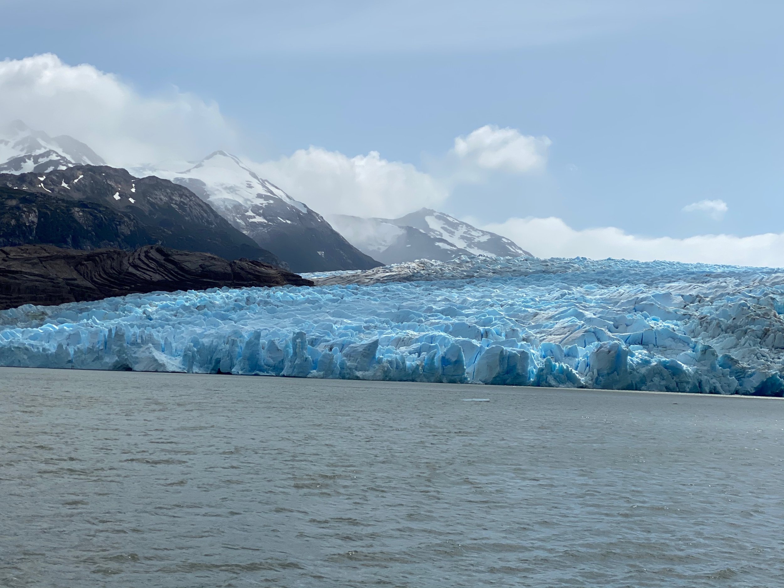 Large blue glacier with mountains in the background and water in the foreground.