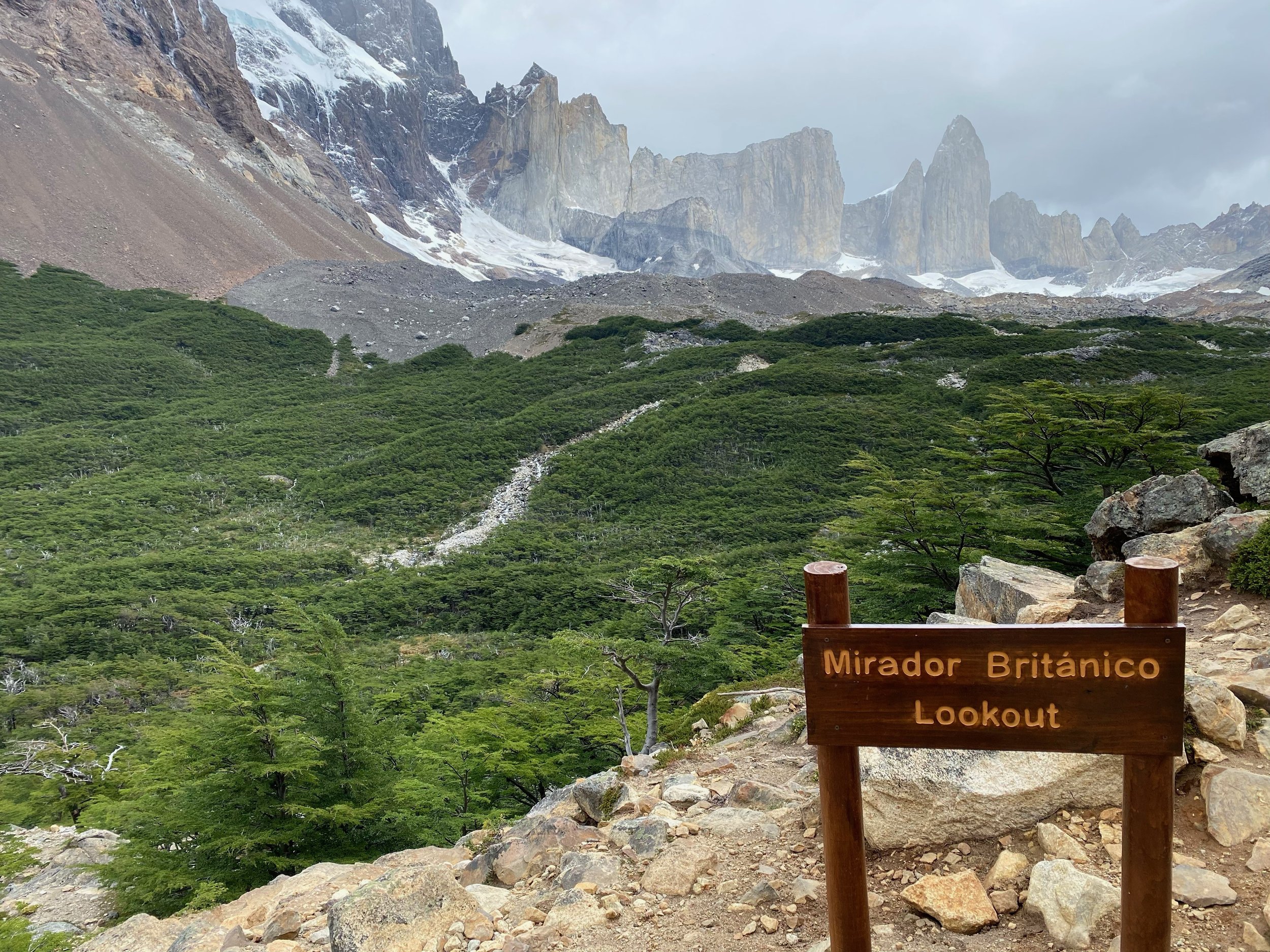 Mirador Británico lookout sign with view of rocky mountains, green forest, and cloudy sky in background.