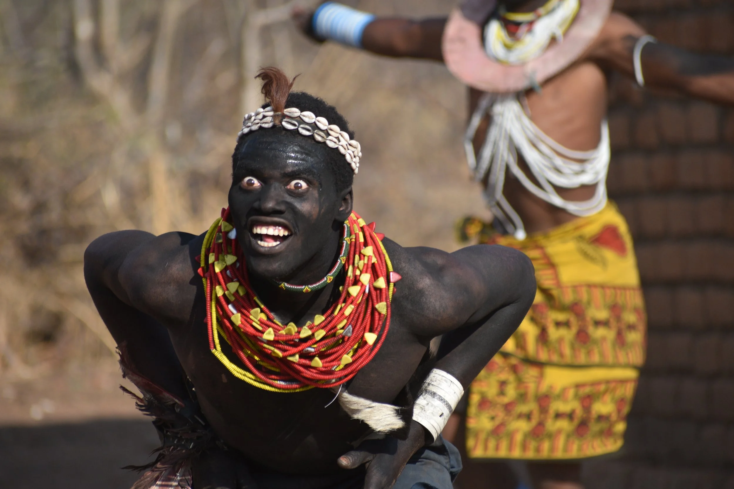 A man with dark skin painted black, wearing multiple colorful beaded necklaces and a headband made of shells, making a dramatic facial expression with wide eyes and an open mouth. In the background, another person is partially visible wearing traditi