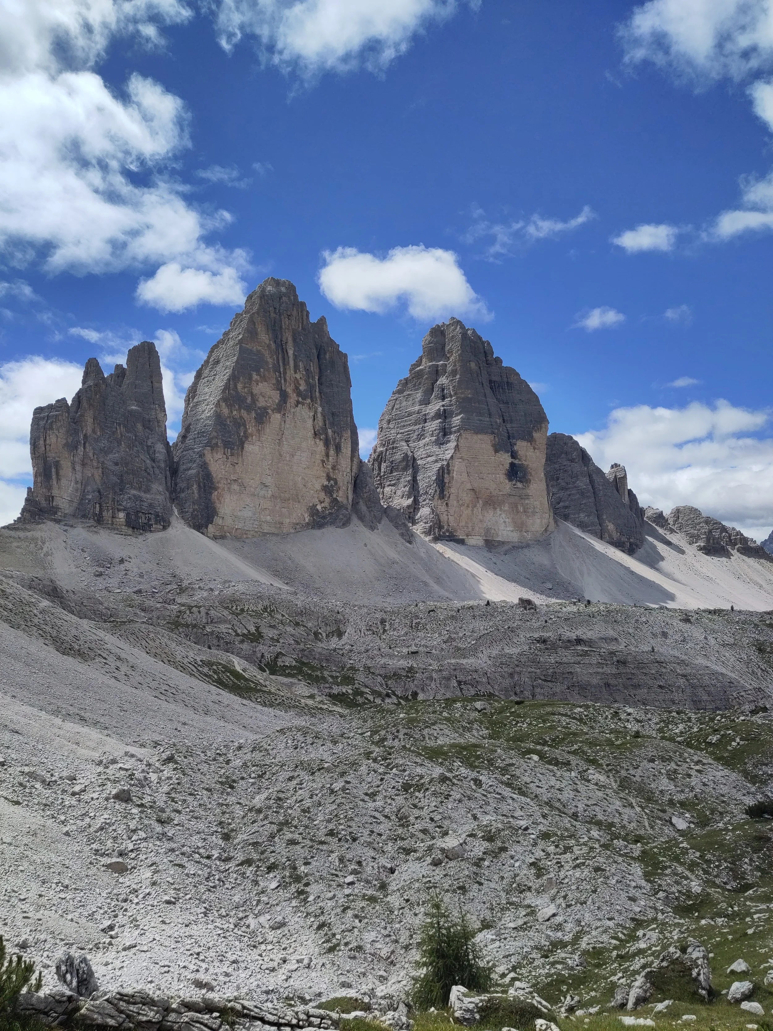 Three towering rock peaks in a mountain landscape with a partly cloudy blue sky and grassy, rocky foreground.