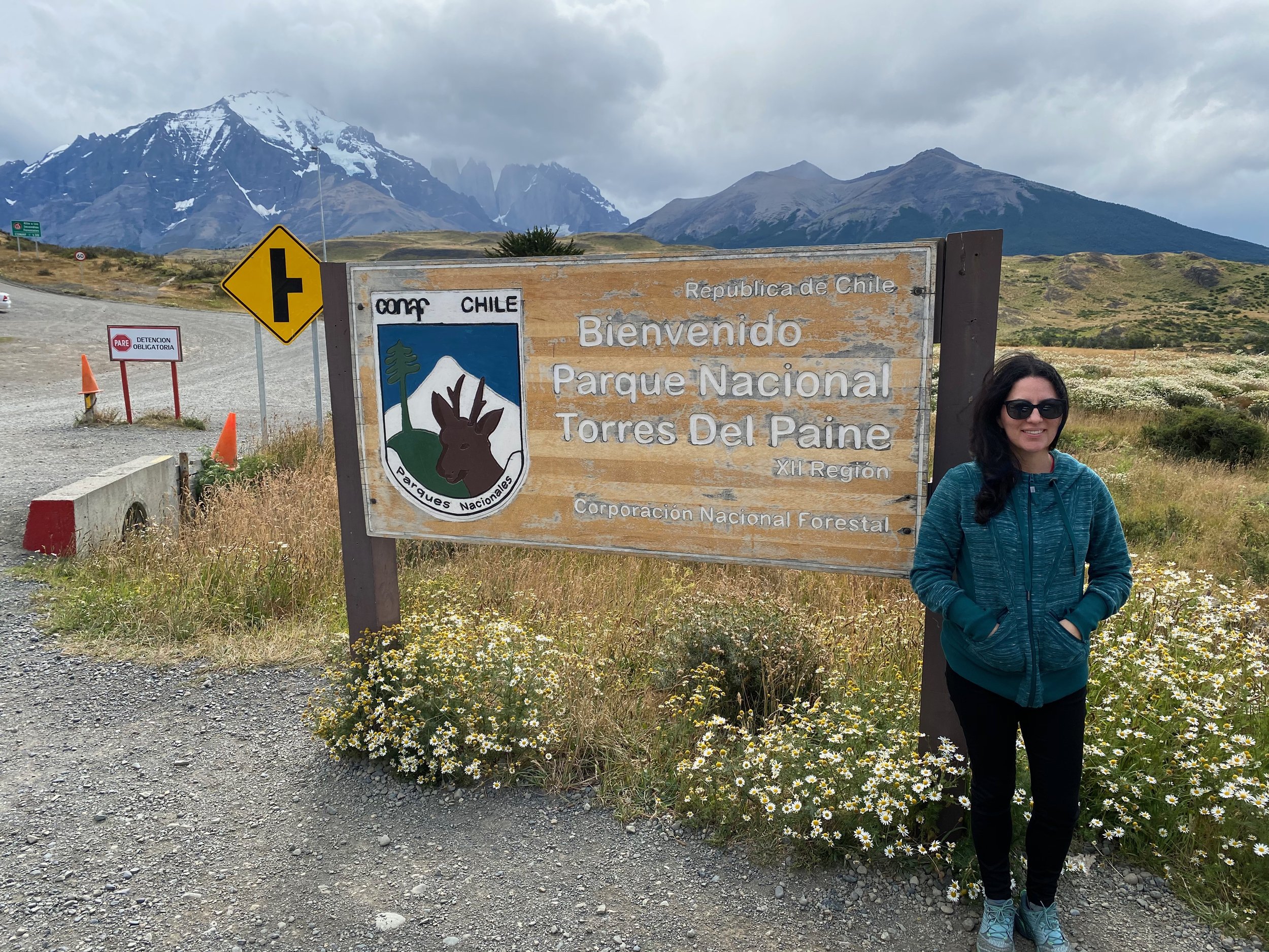 Woman standing next to a sign at the entrance of Torres del Paine National Park, Chile, with mountains in the background.