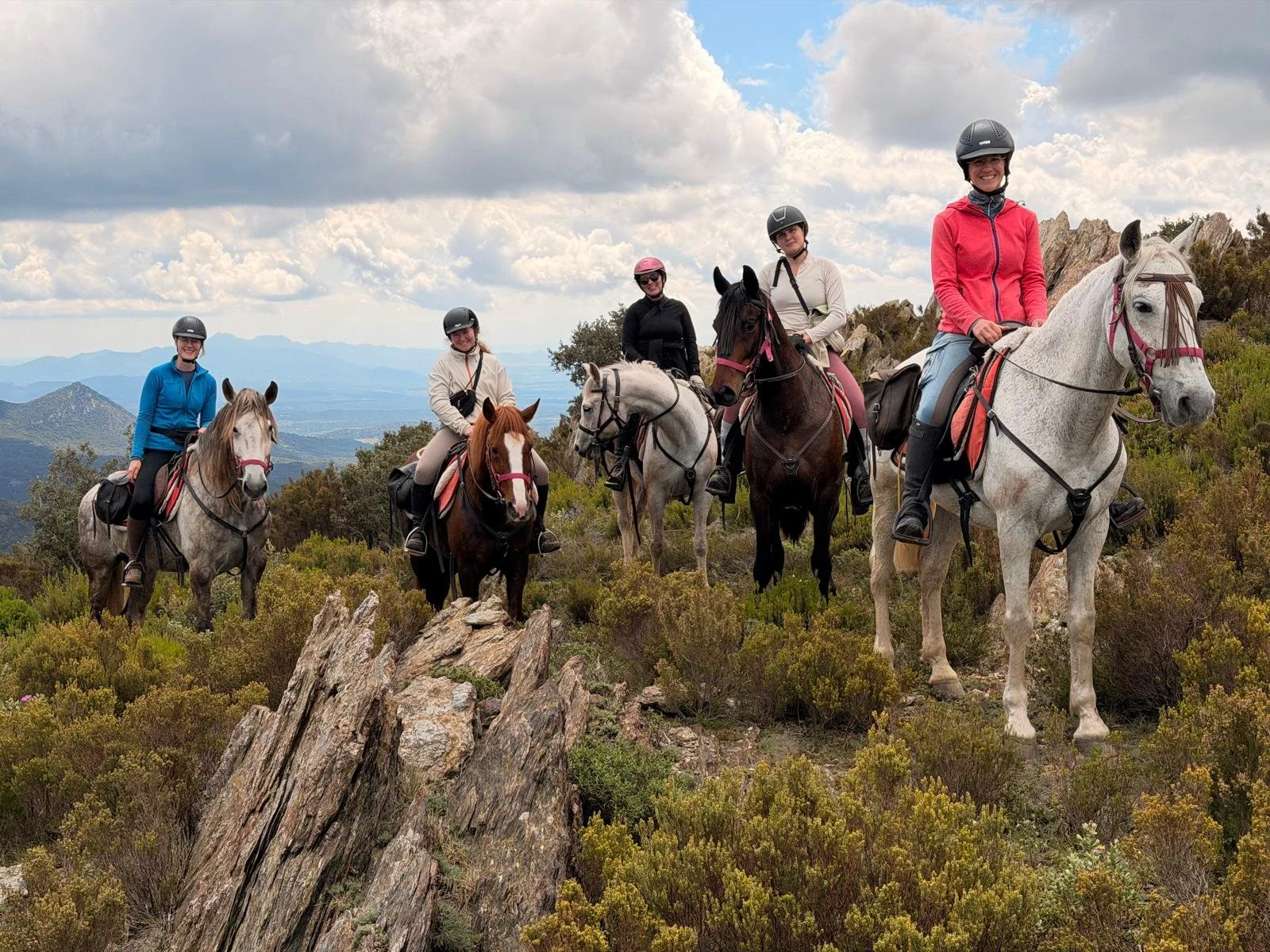 Five women riding horses along a mountain trail, dressed in riding gear and helmets, overlooking a scenic landscape with mountains, clouds, and green shrubbery.