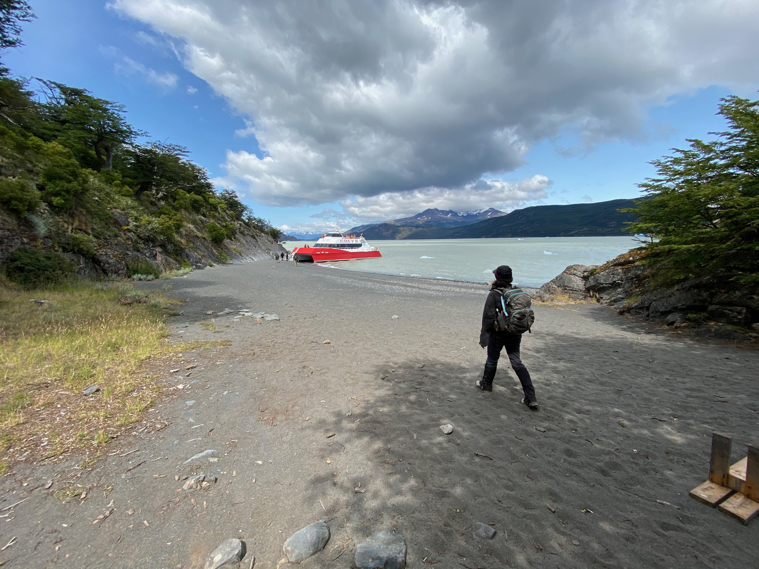 Person walking on a rocky beach towards a red and white boat near the shore, surrounded by green hills and a cloudy sky.