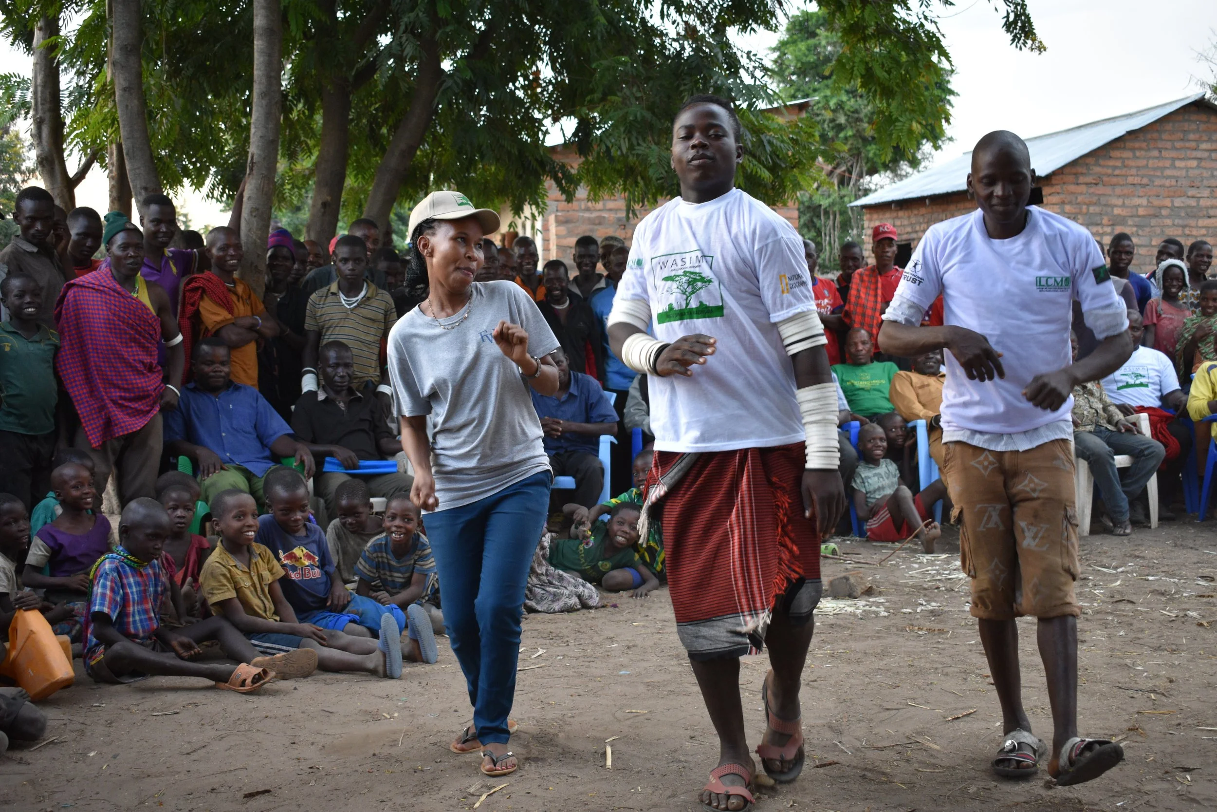 Three people dancing outdoors in front of an audience of children and adults seated and watching, with trees and buildings in the background.