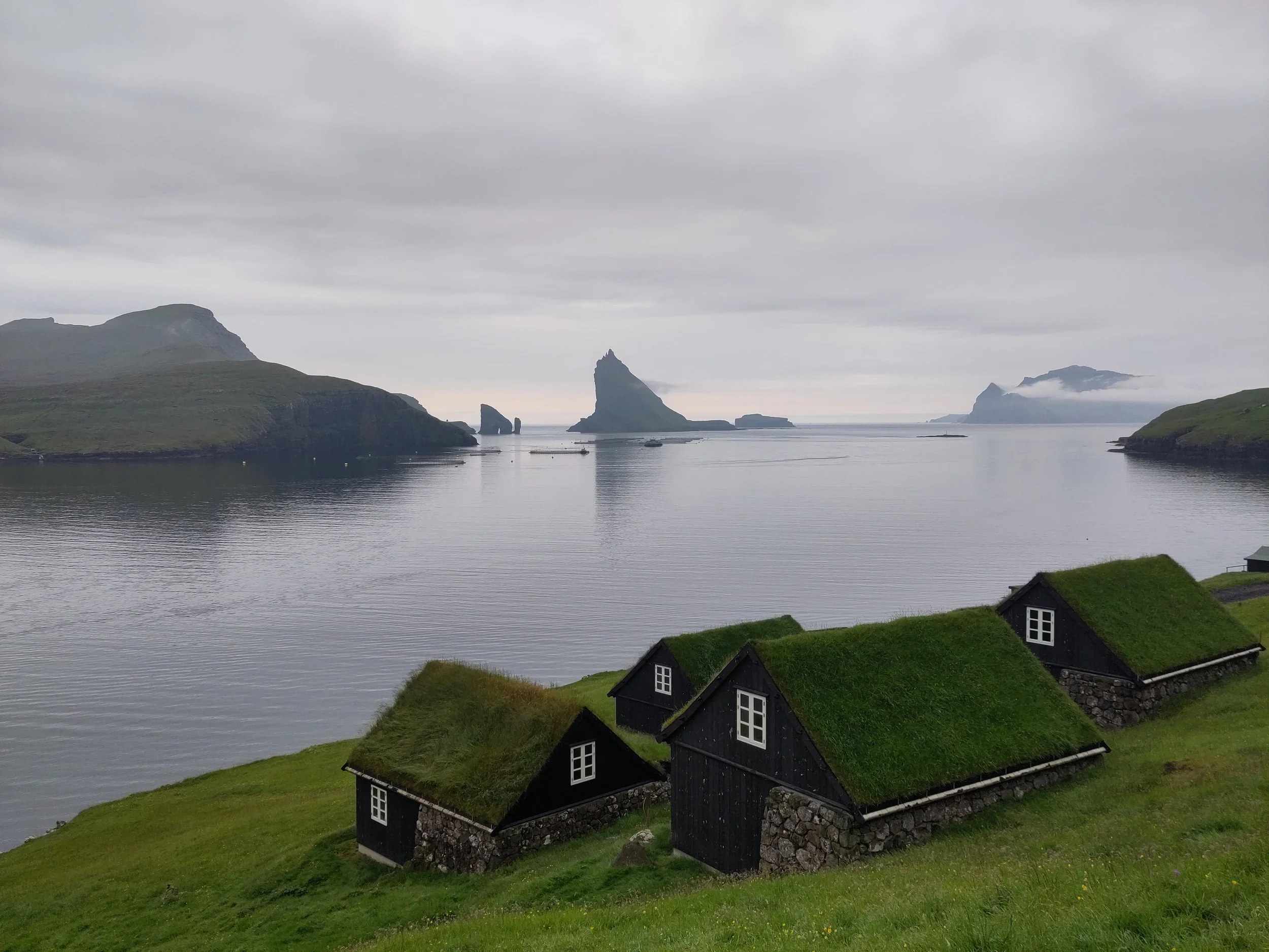 Small houses with green grass roofs on a green hillside overlooking a calm body of water with rocky islands and mountains in the distance under cloudy sky.