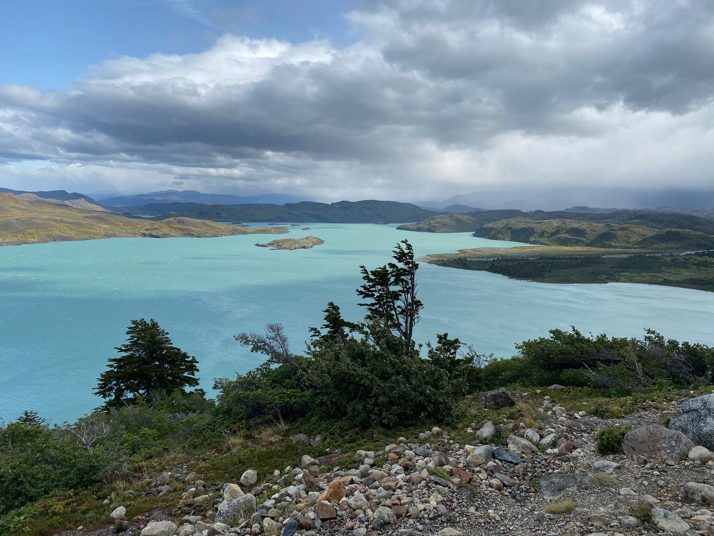 Scenic view of a turquoise lake surrounded by rocky shoreline and hills with a cloudy sky overhead.