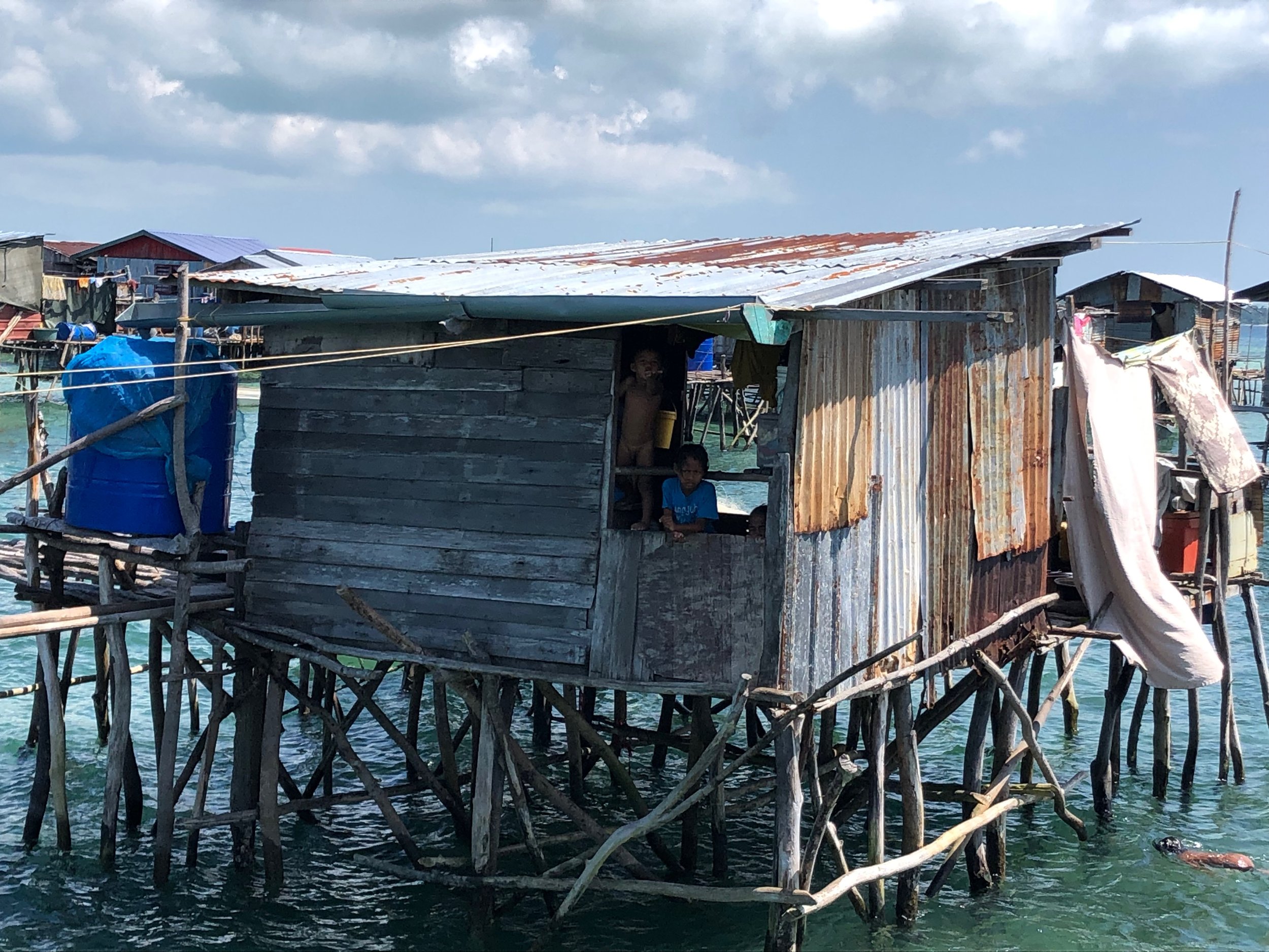 Wooden stilt house over water with children peering from doorway, surrounded by rustic materials and blue sky.