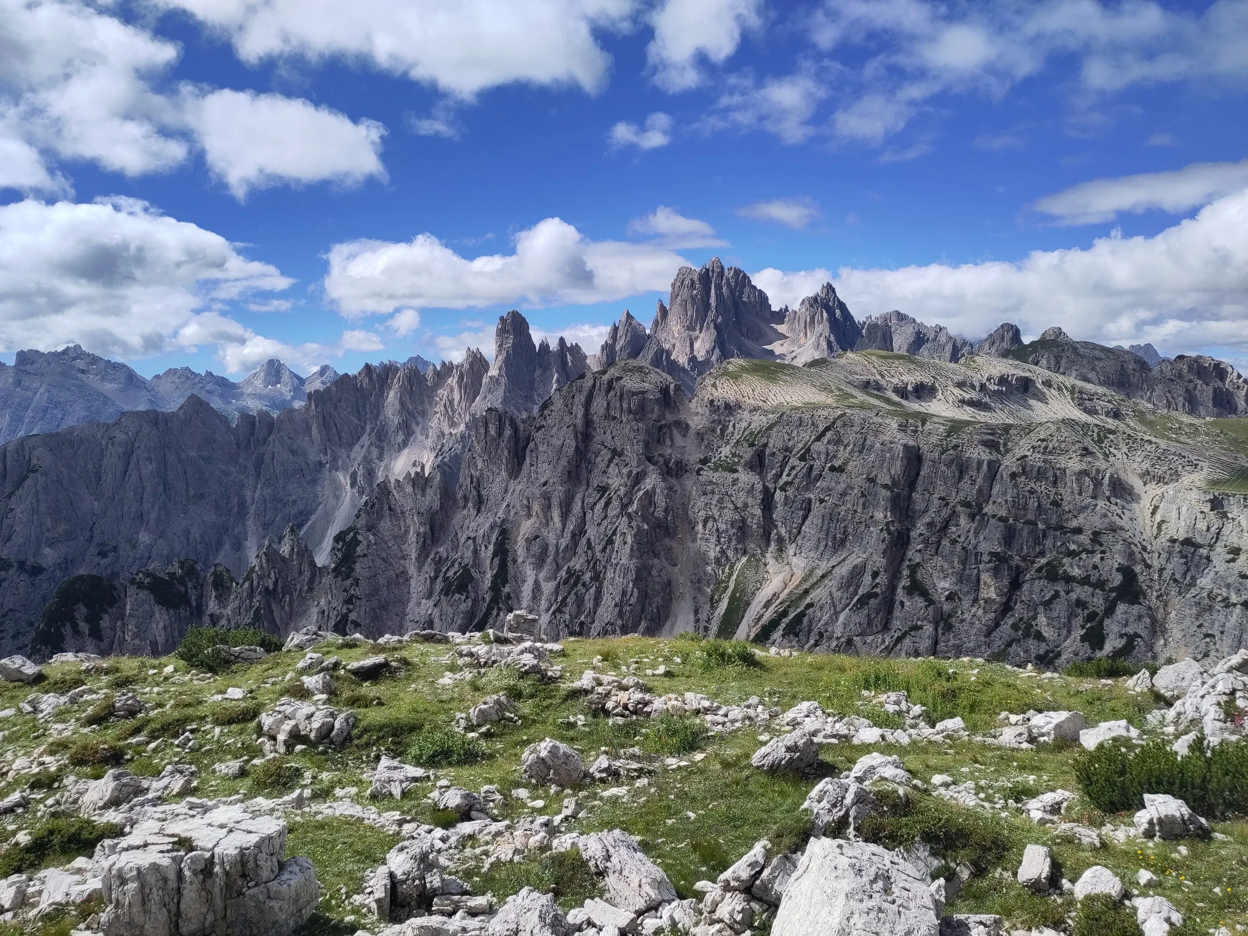 Mountain range with sharp peaks under a partly cloudy sky, with a foreground of rocky and grassy terrain.