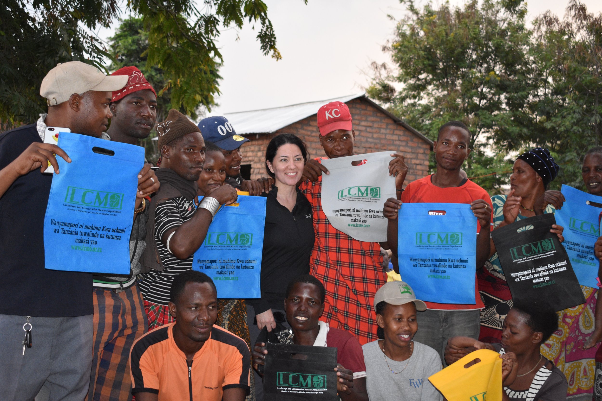 Group of men and women holding blue and black eco-bags, some wearing hats, standing outdoors with trees and a brick house in the background, smiling and posing for the photo.