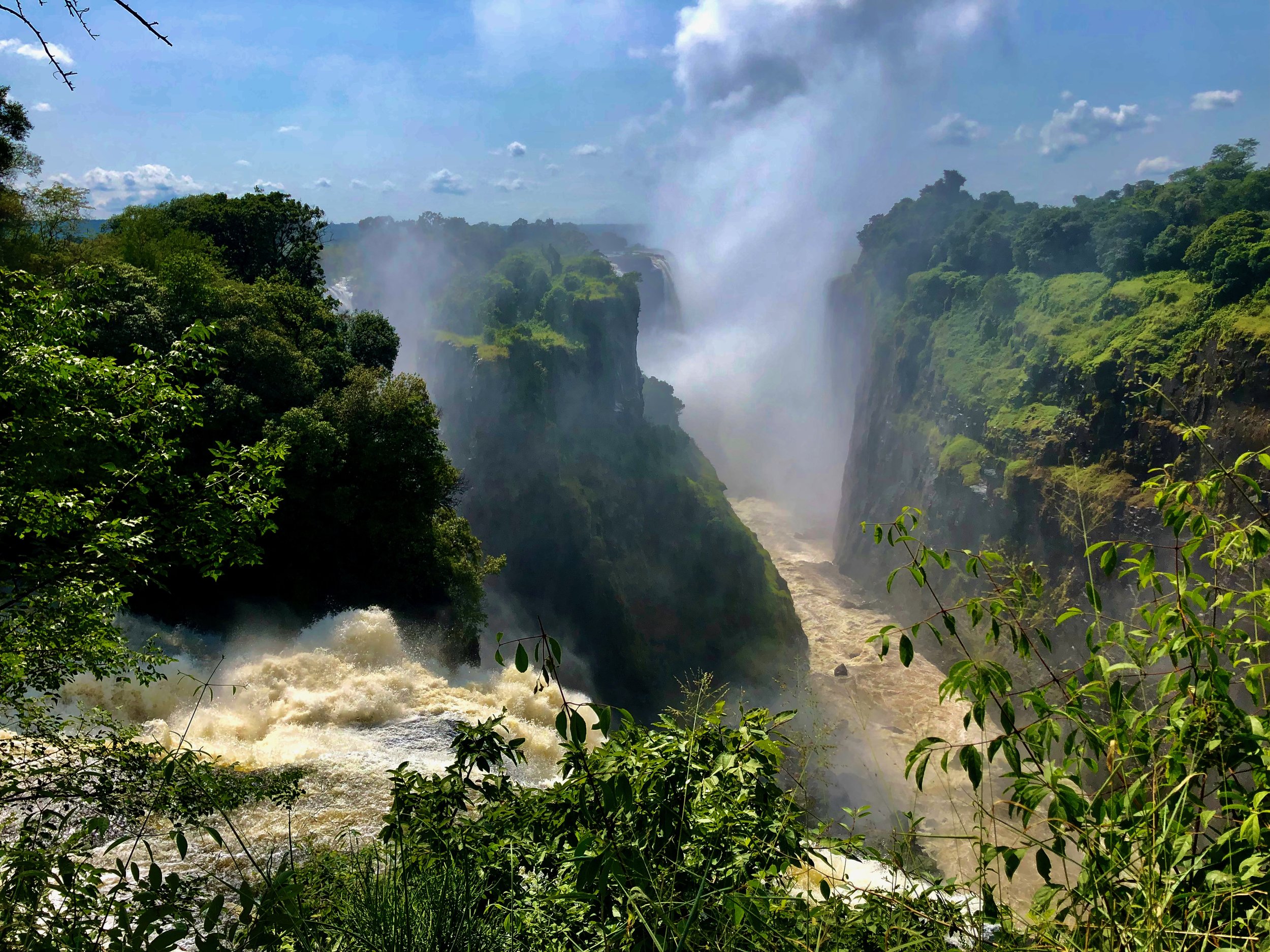 View of a large waterfall in a lush green jungle, with water cascading down cliffs and mist rising from the rushing water below.